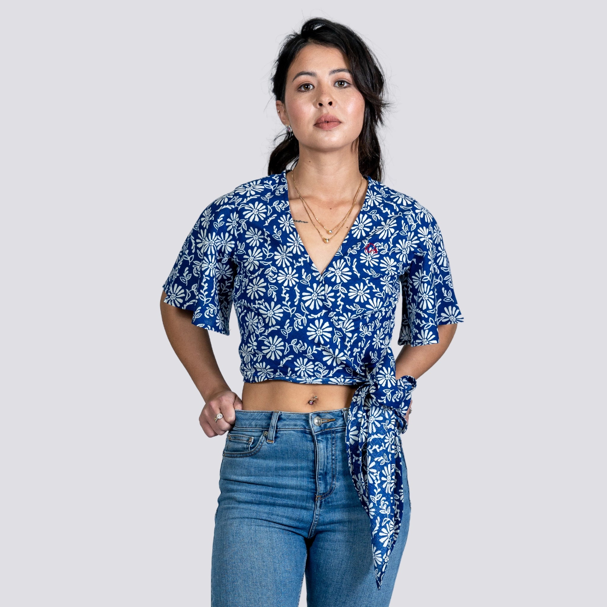 Young woman in a SereneBloom Women's Eco-Friendly Wrap Top - Blue Bliss by Karee and jeans, standing confidently with one hand on her hip, against a light gray background.