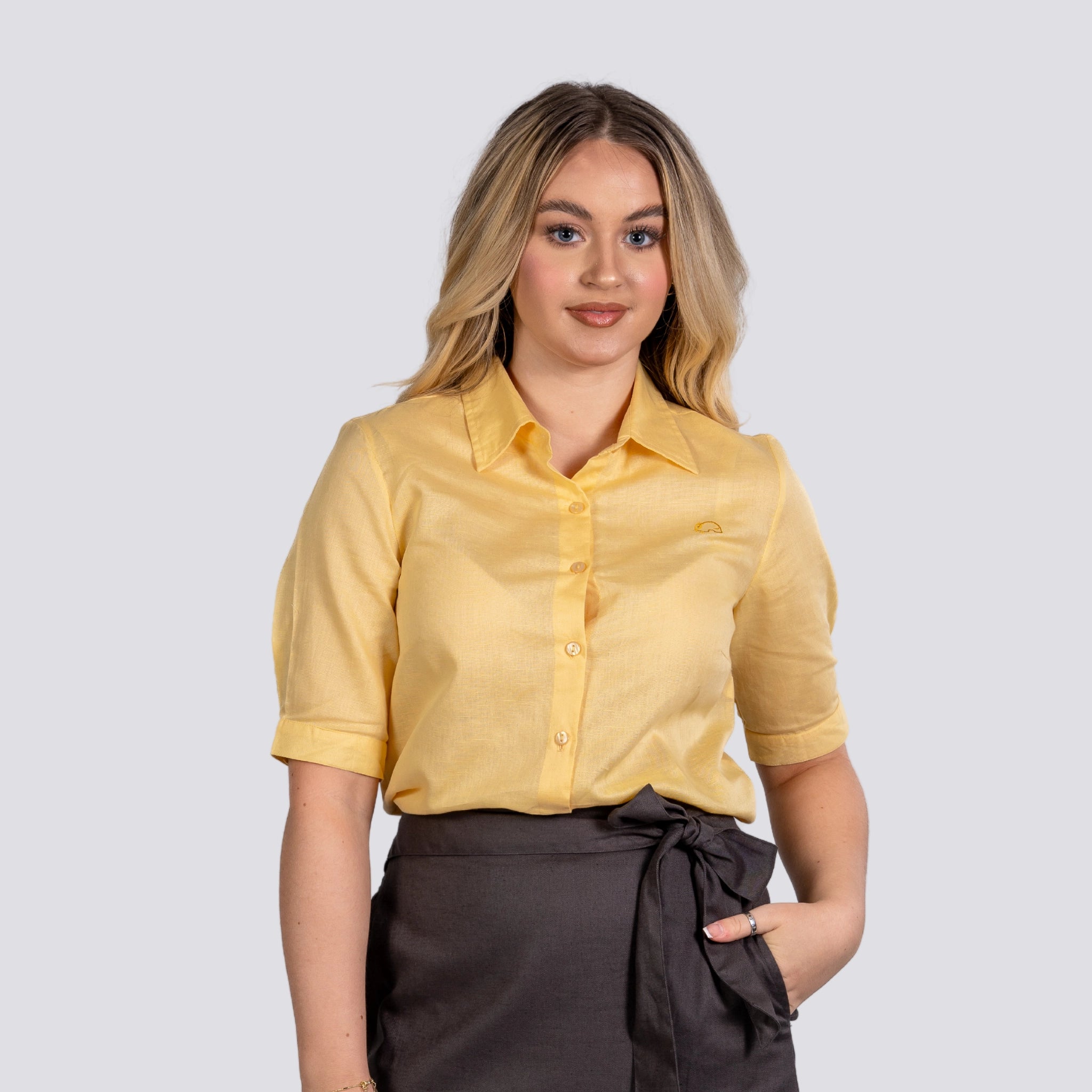 A young woman wearing a Karee Sunlit Breeze Yellow Linen Cotton Shirt For Women and brown skirt, standing with her hand on her hip, against a light gray background.