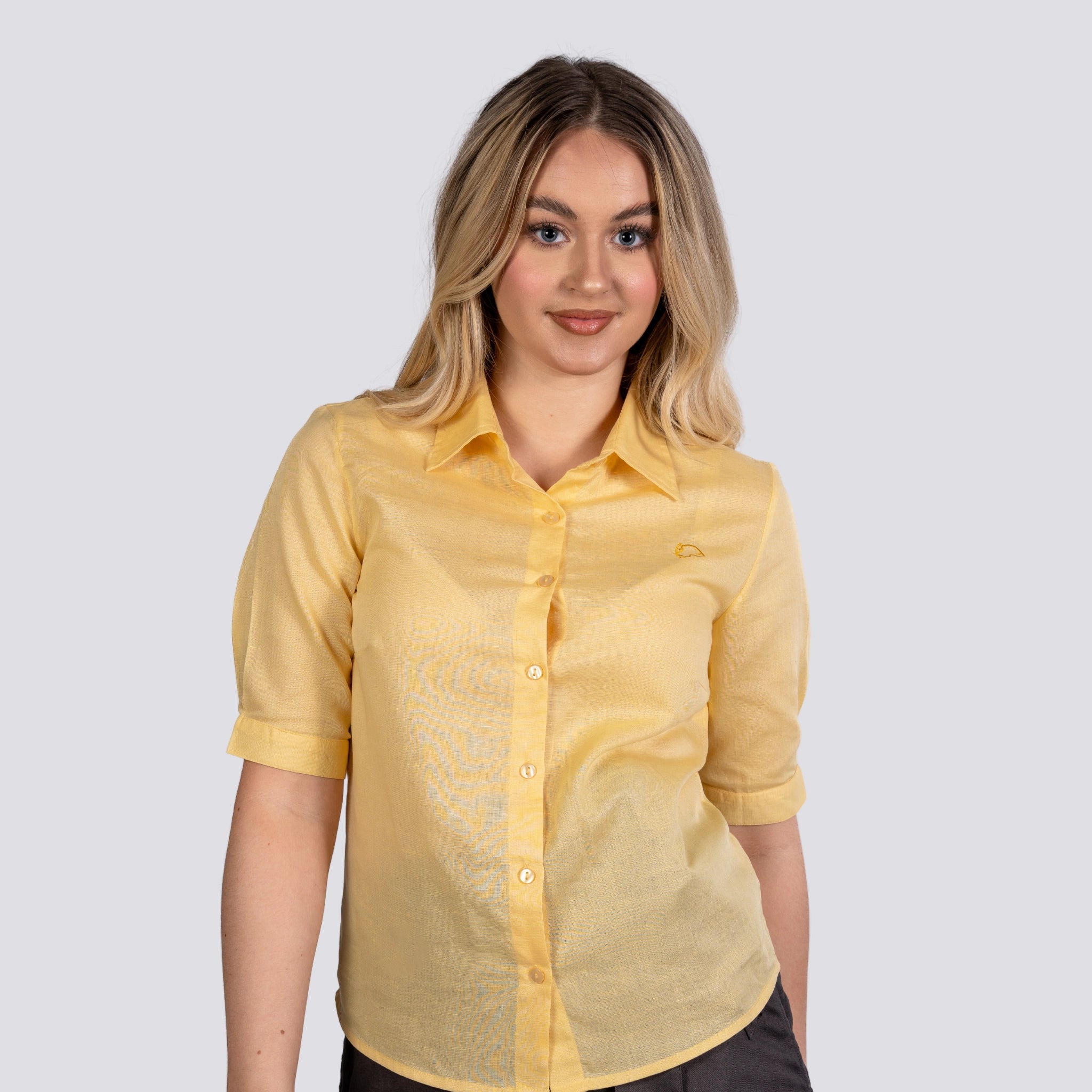 A young woman sporting a Karee Sunlit Breeze Yellow Linen Cotton Shirt For Women, standing against a gray background, smiling slightly at the camera.