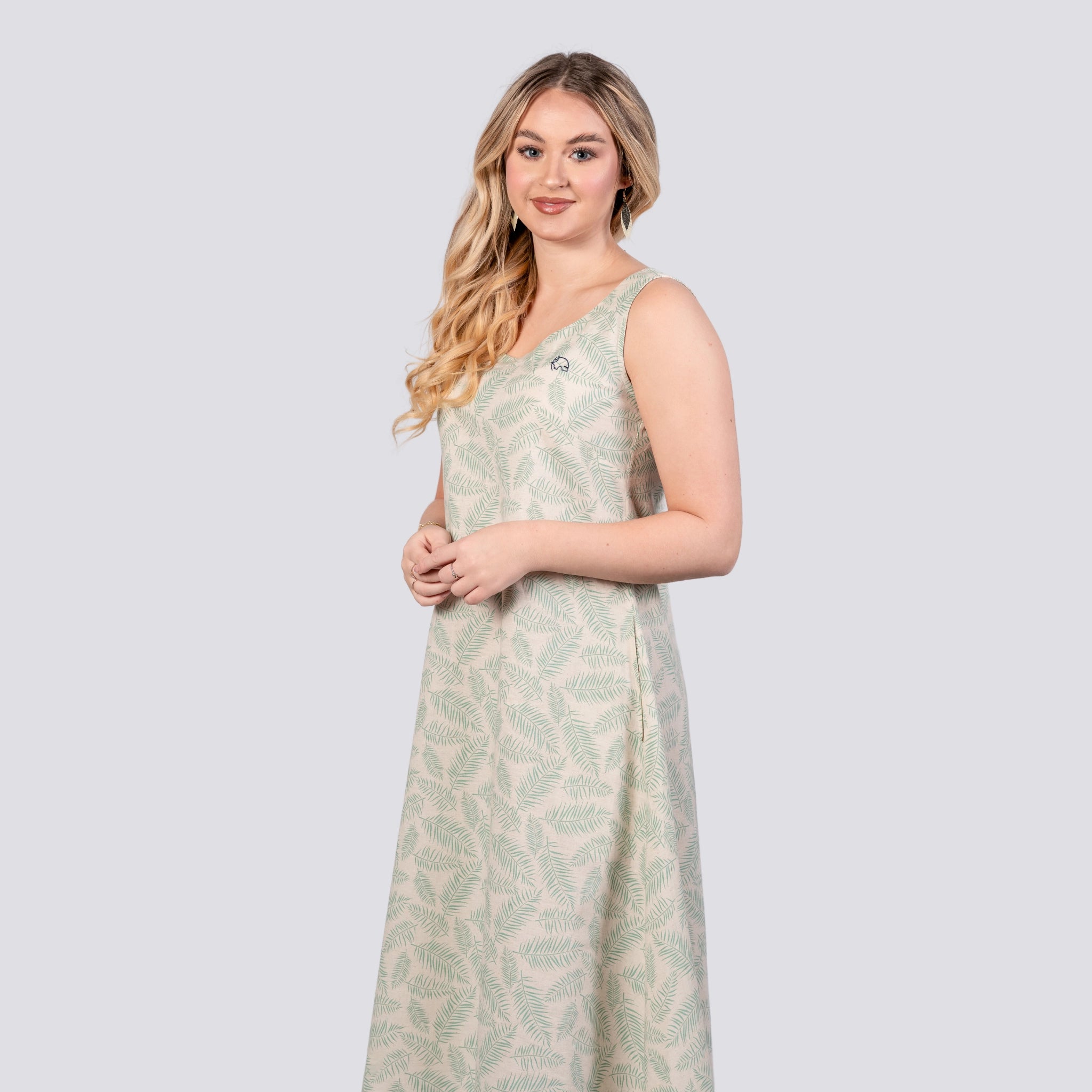 A woman with long blonde hair wearing a light green patterned Eco-Luxe Look: Sustainable Mystic Breeze Linen Hi-Lo Midi Dress for Women by Karee stands against a plain background. The dress, made of a linen cotton blend, features a high low hemline. She faces the camera with her hands clasped in front of her.