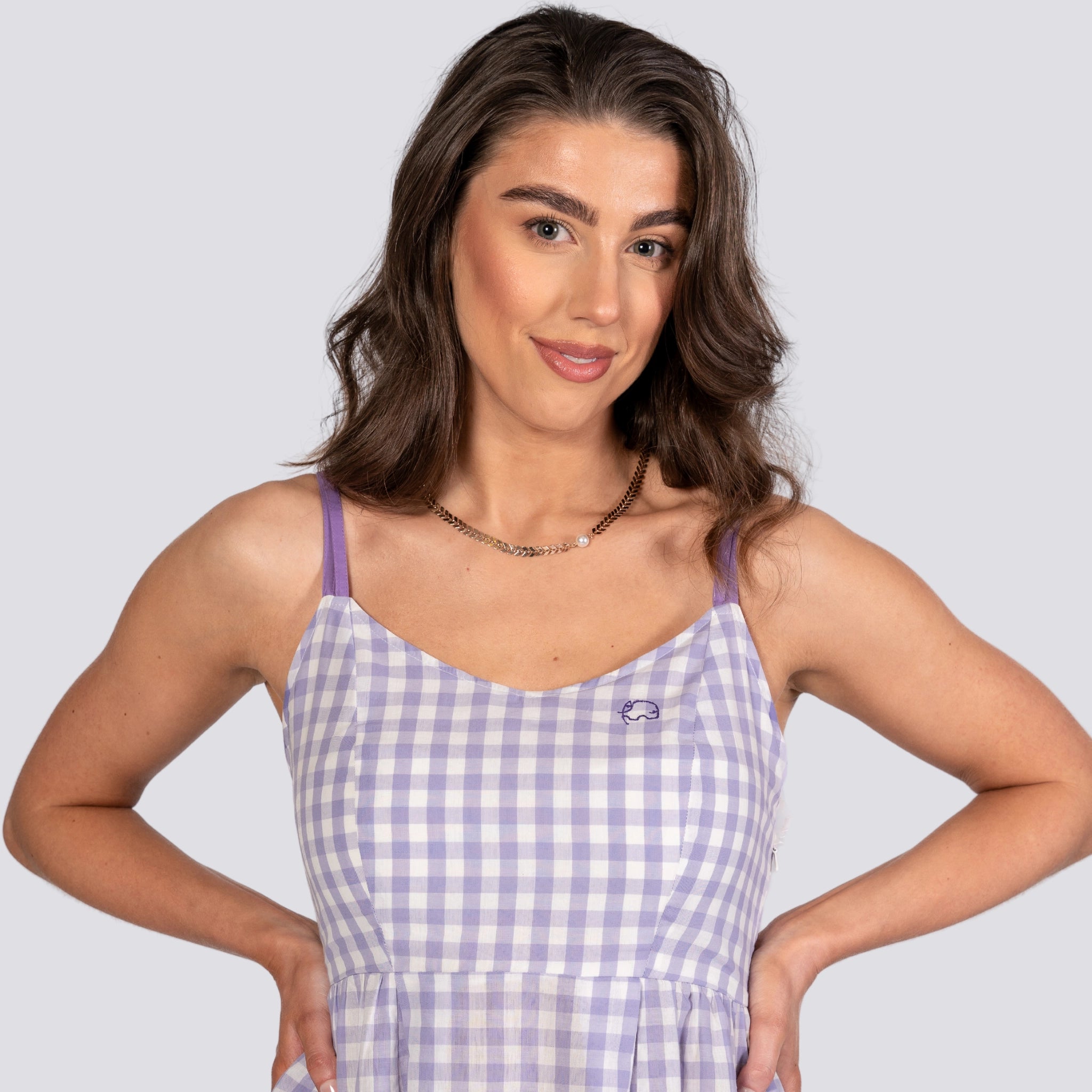 A woman stands with her hands on her hips, wearing the Karee Effortless Summer Style: Sustainable Lavender Gingham Mini Dress with a small whale logo. Crafted from breathable cotton, the limited-edition dress complements her long wavy hair as she smiles slightly.