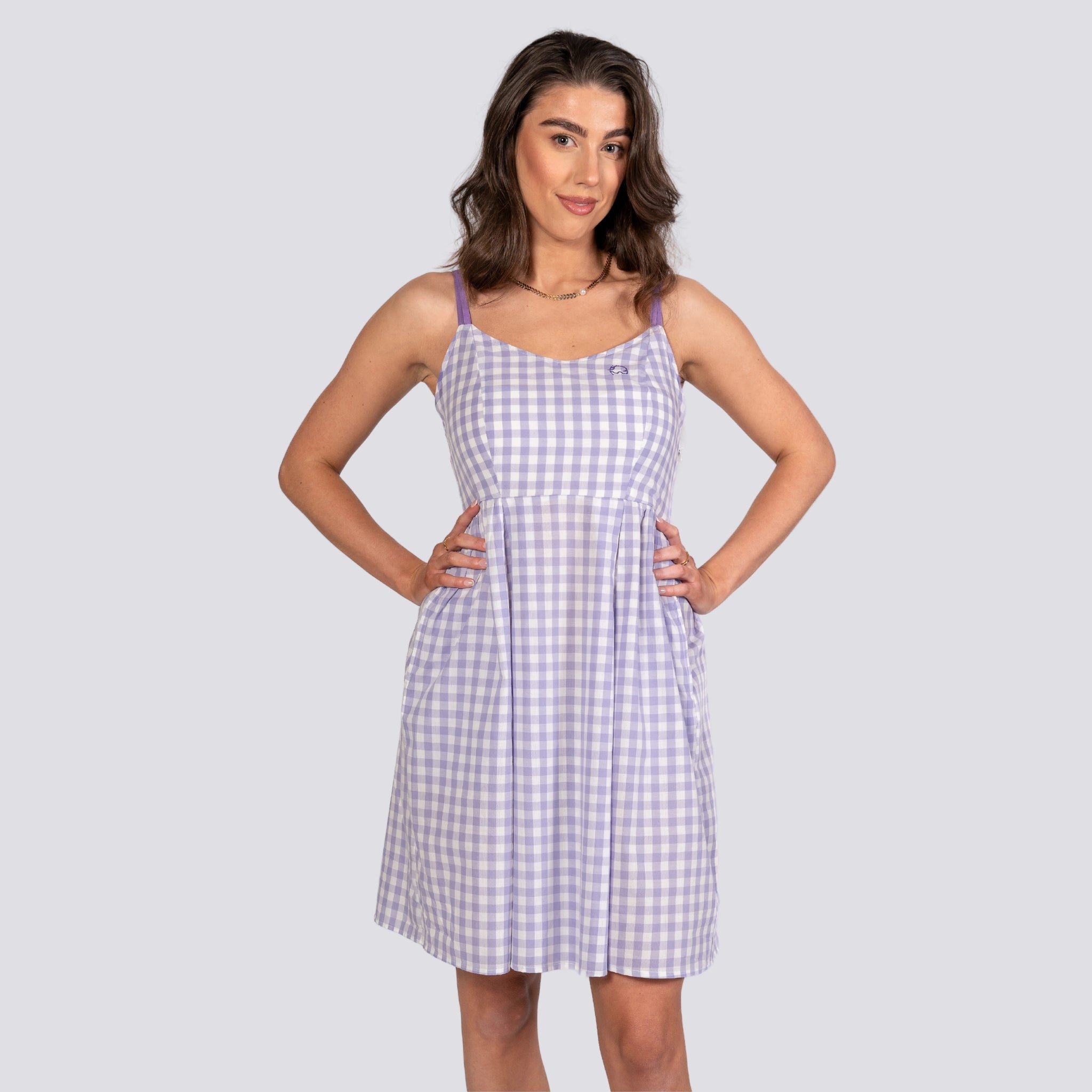 A woman stands with hands on hips, wearing a sleeveless, knee-length Effortless Summer Style: Sustainable Lavender Gingham Mini Dress by Karee in shades of white and purple.