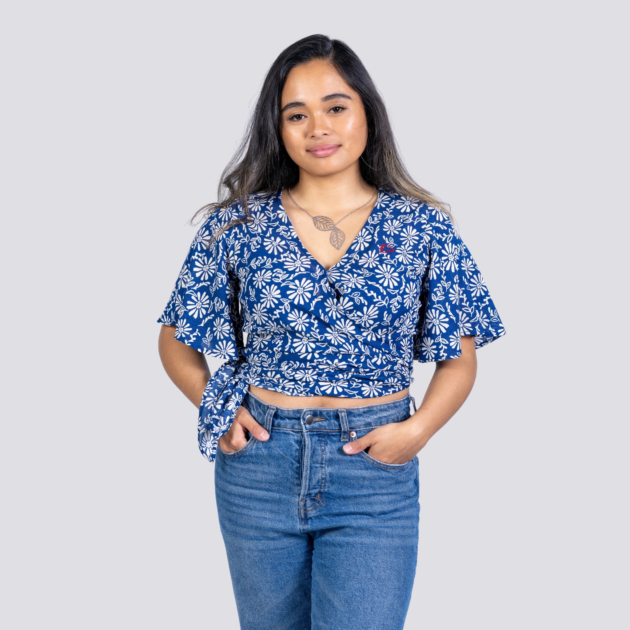 A woman wearing a Karee SereneBloom Women's Eco-Friendly Wrap Top - Blue Bliss and jeans stands with her hands on hips against a grey background.