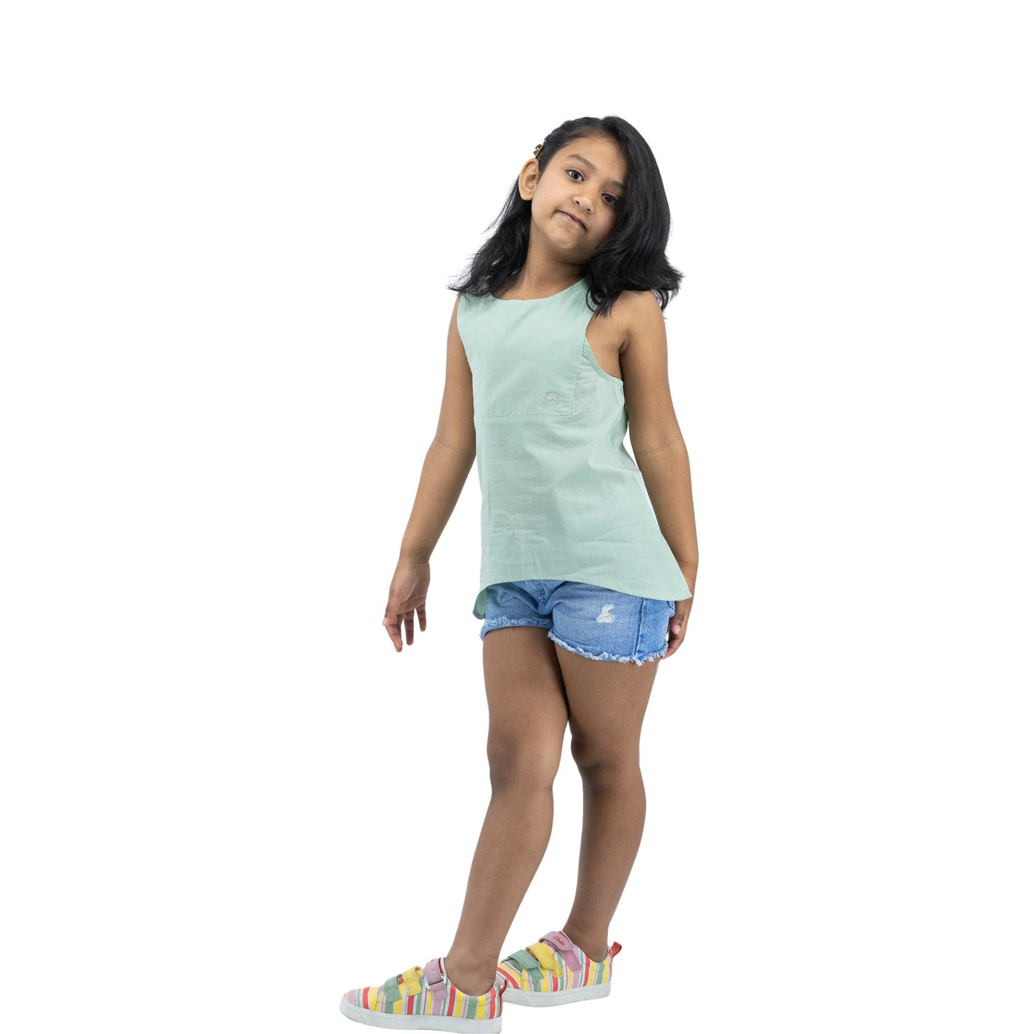 A young girl in a Karee Smoke Green Cotton Bib Neck Top for kids and denim shorts stands confidently against a white background.