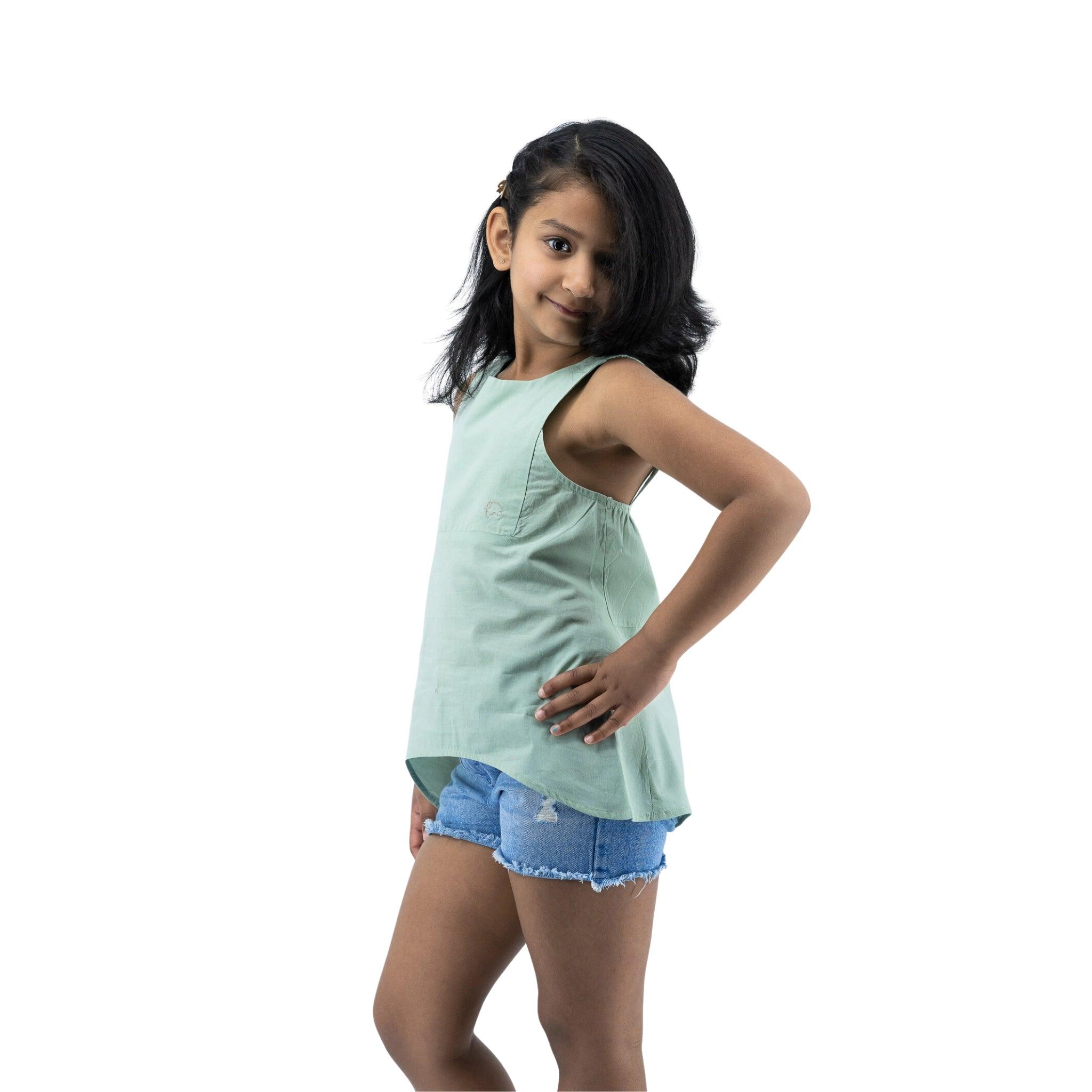 A young girl in a Karee Smoke Green Cotton Bib Neck Top and denim shorts stands smiling with her hands on her hips, isolated on a white background.