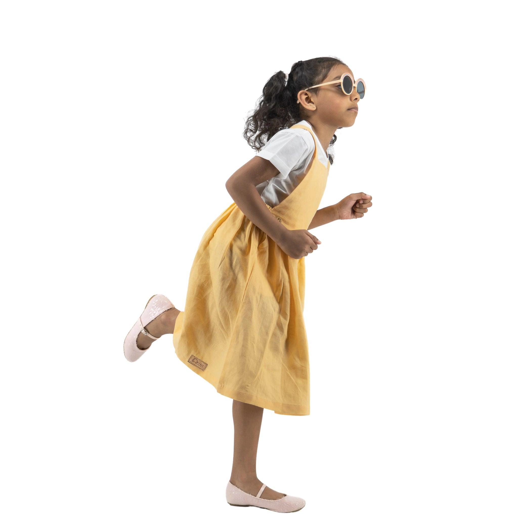 A young girl in a Karee Golden Fleece Linen Pinafore and white sunglasses playfully running, isolated on a white background.