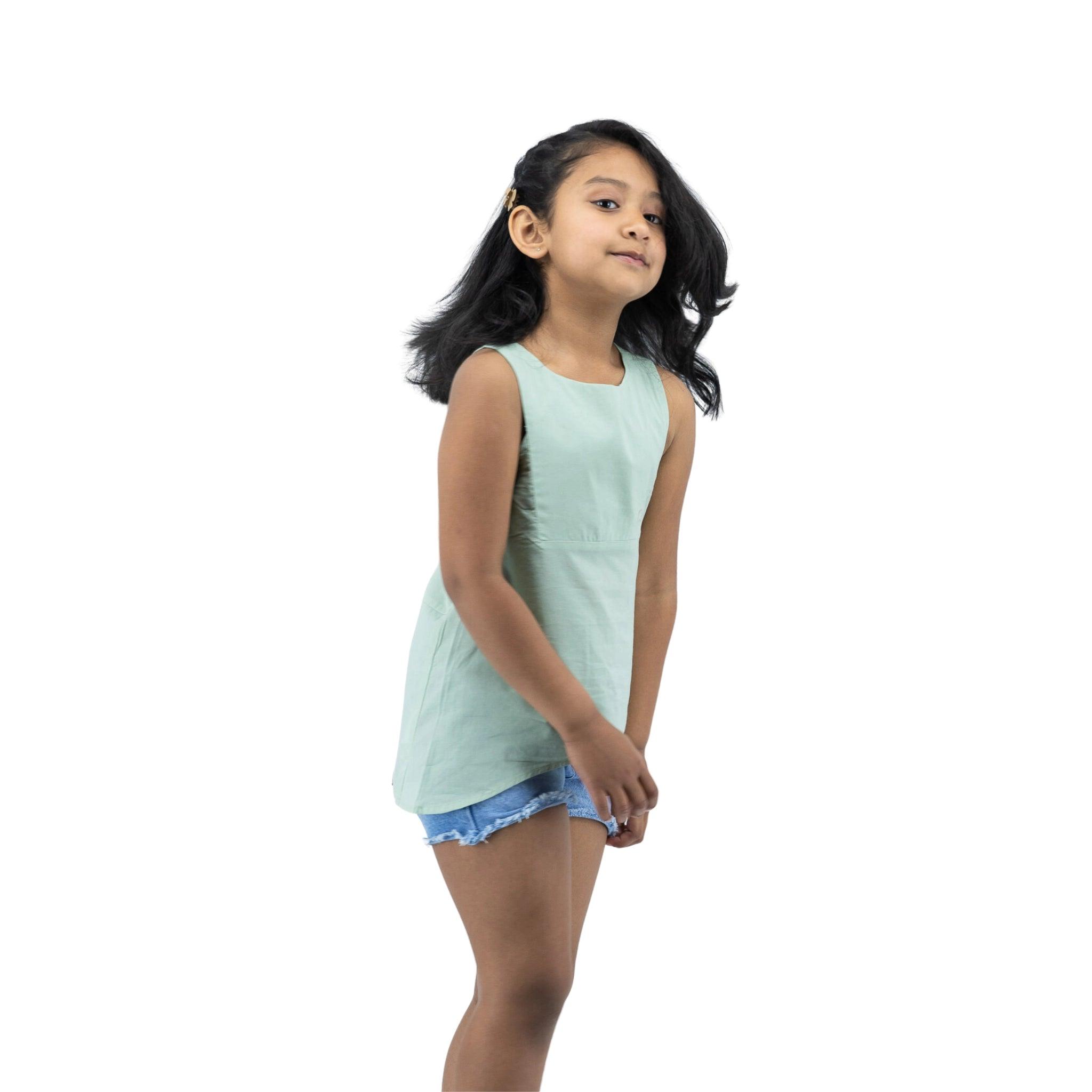 Young girl in a Karee Smoke Green Cotton Bib Neck Top for kids and denim shorts walking to her right, looking away, with a white background.