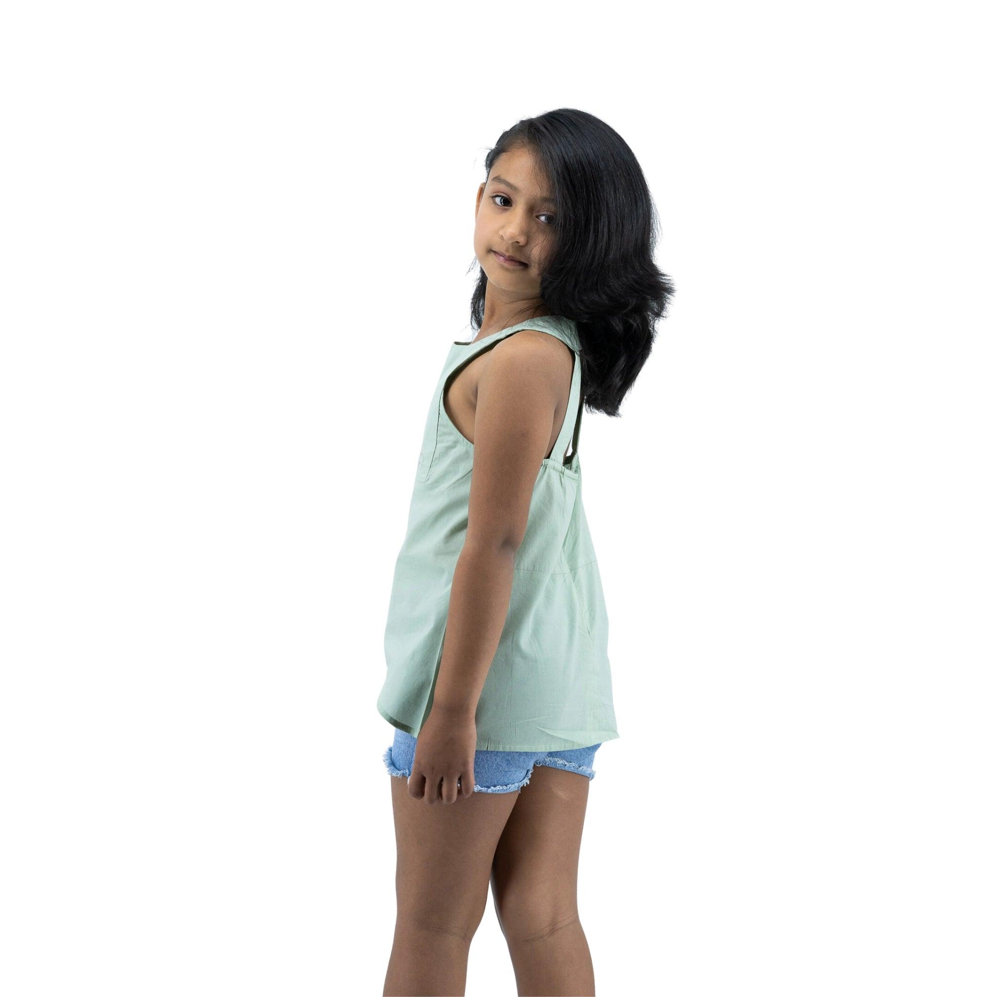 Young girl in a Karee Smoke Green Cotton Bib Neck Top for kids and denim shorts standing sideways, looking over her shoulder, against a white background.