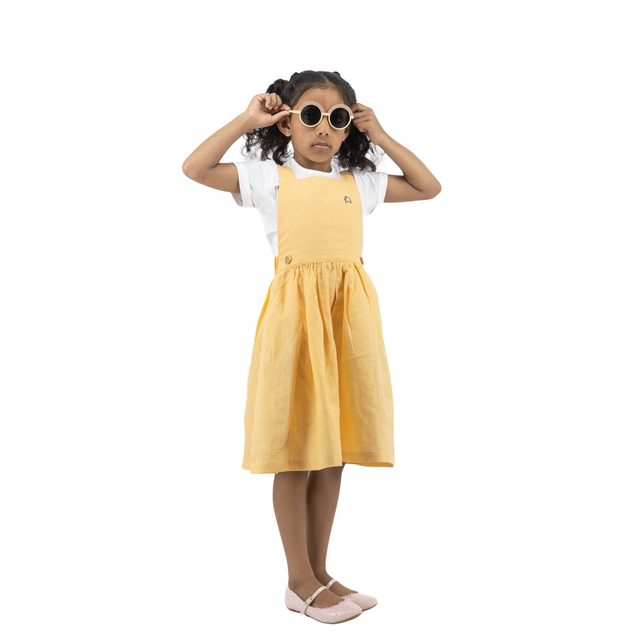 Young girl in a Karee Golden Fleece Linen Pinafore dress and white shirt adjusting oversized sunglasses, standing against a white background.