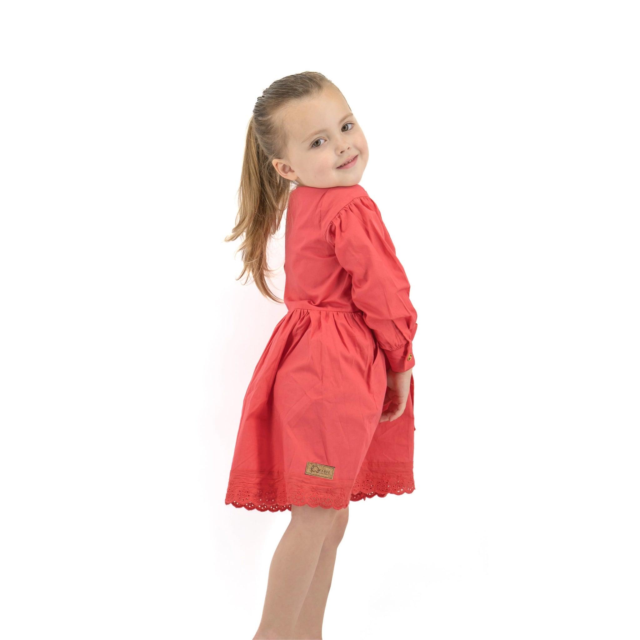 A young girl in a Karee red long puff sleeve cotton dress stands smiling, looking over her shoulder against a white background.