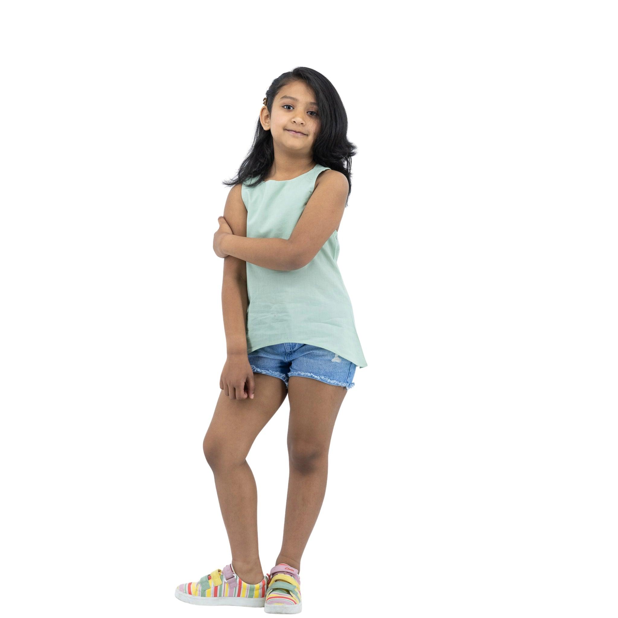 Young girl standing with arms crossed, wearing a Karee Smoke Green Cotton Bib Neck Top for kids, denim shorts, and colorful sneakers, isolated on a white background.