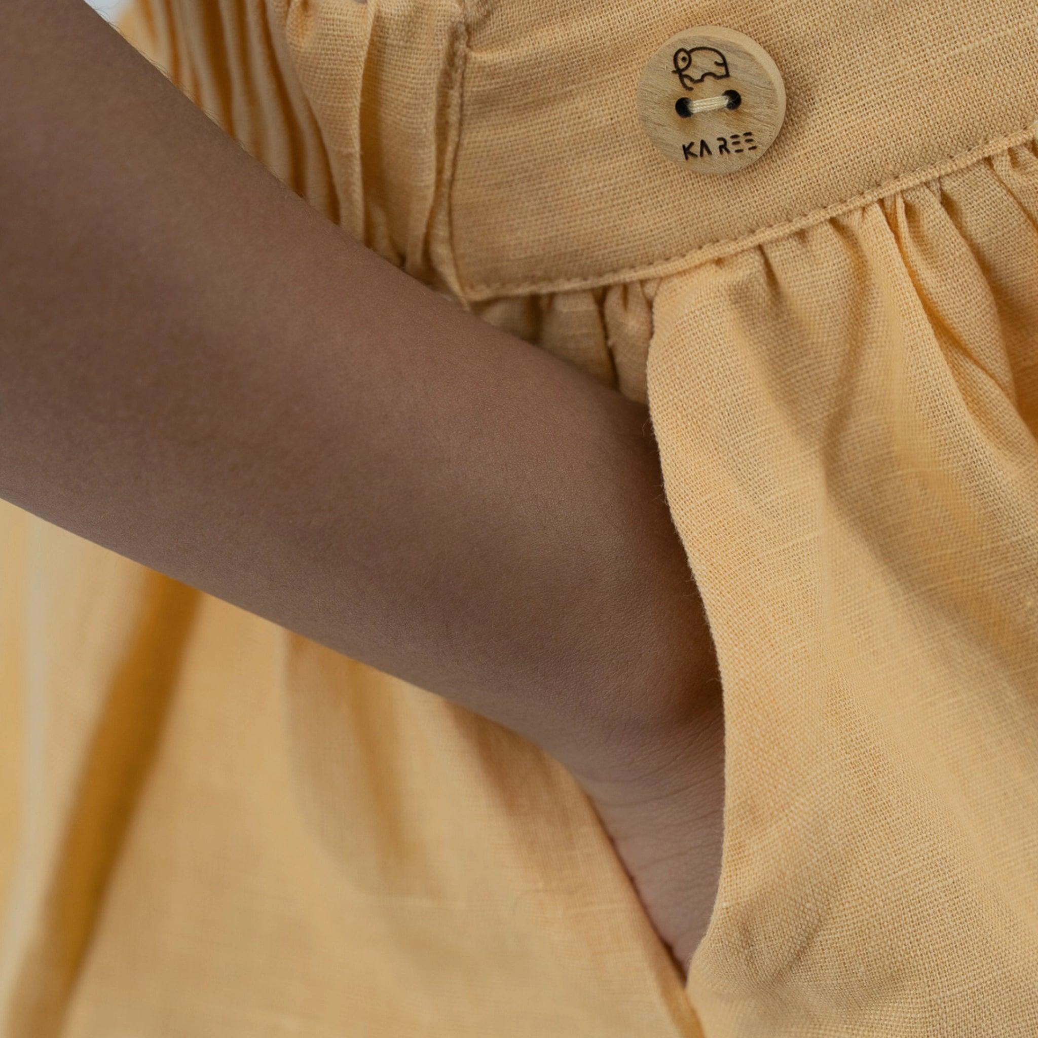 Close-up of a person wearing a yellow Karee Golden Fleece Linen Pinafore dress with a wooden button labeled "kare" at the pocket.