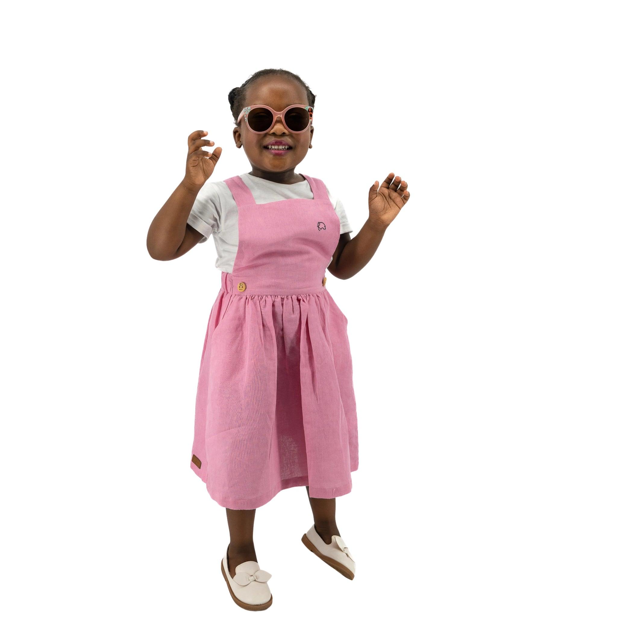Young girl in a pink Karee Cashmere Rose Linen pinafore, smiling and waving, isolated on a white background.