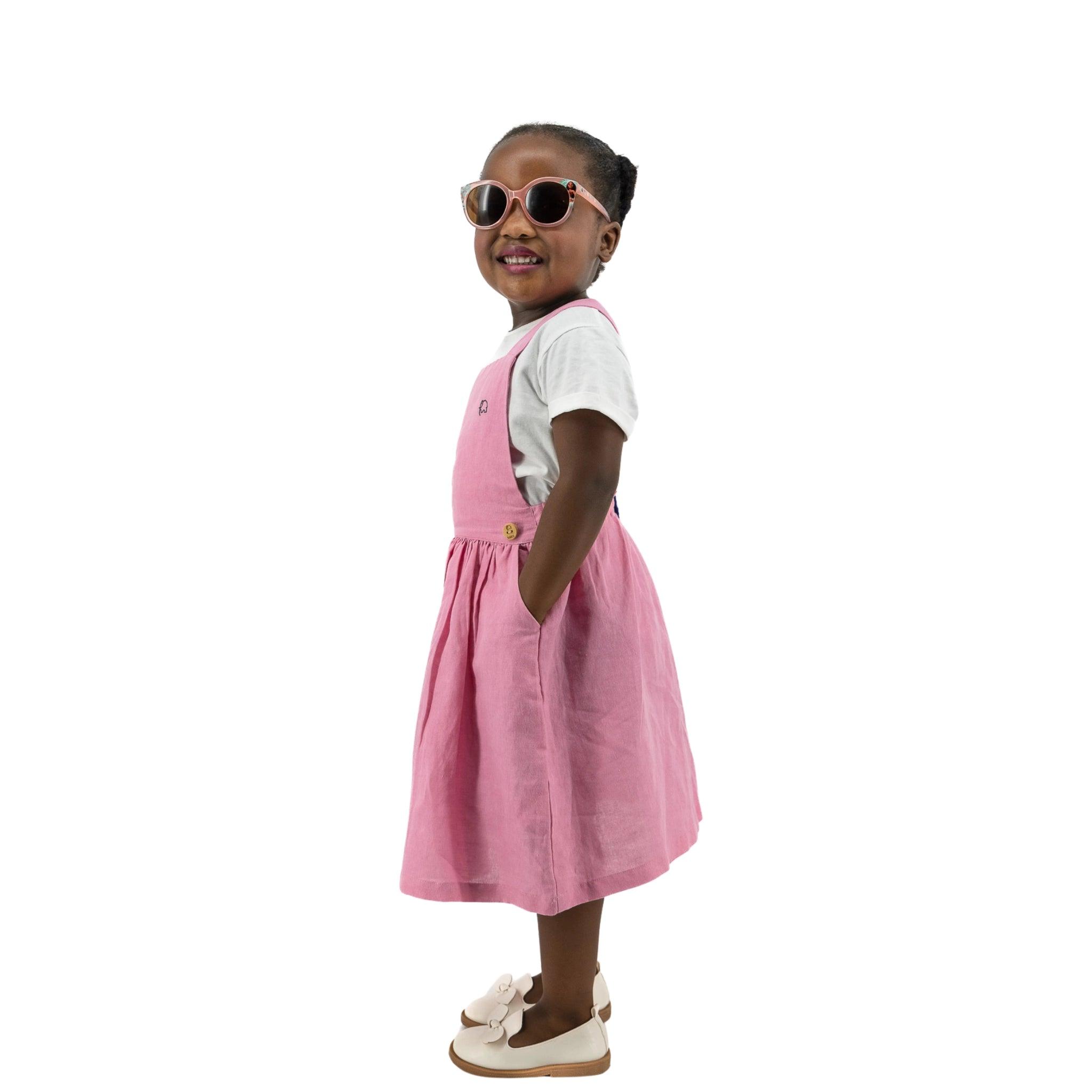 Young girl in a Karee Cashmere Rose Linen Pinafore and sunglasses, standing confidently with hands on hips, isolated on a white background.