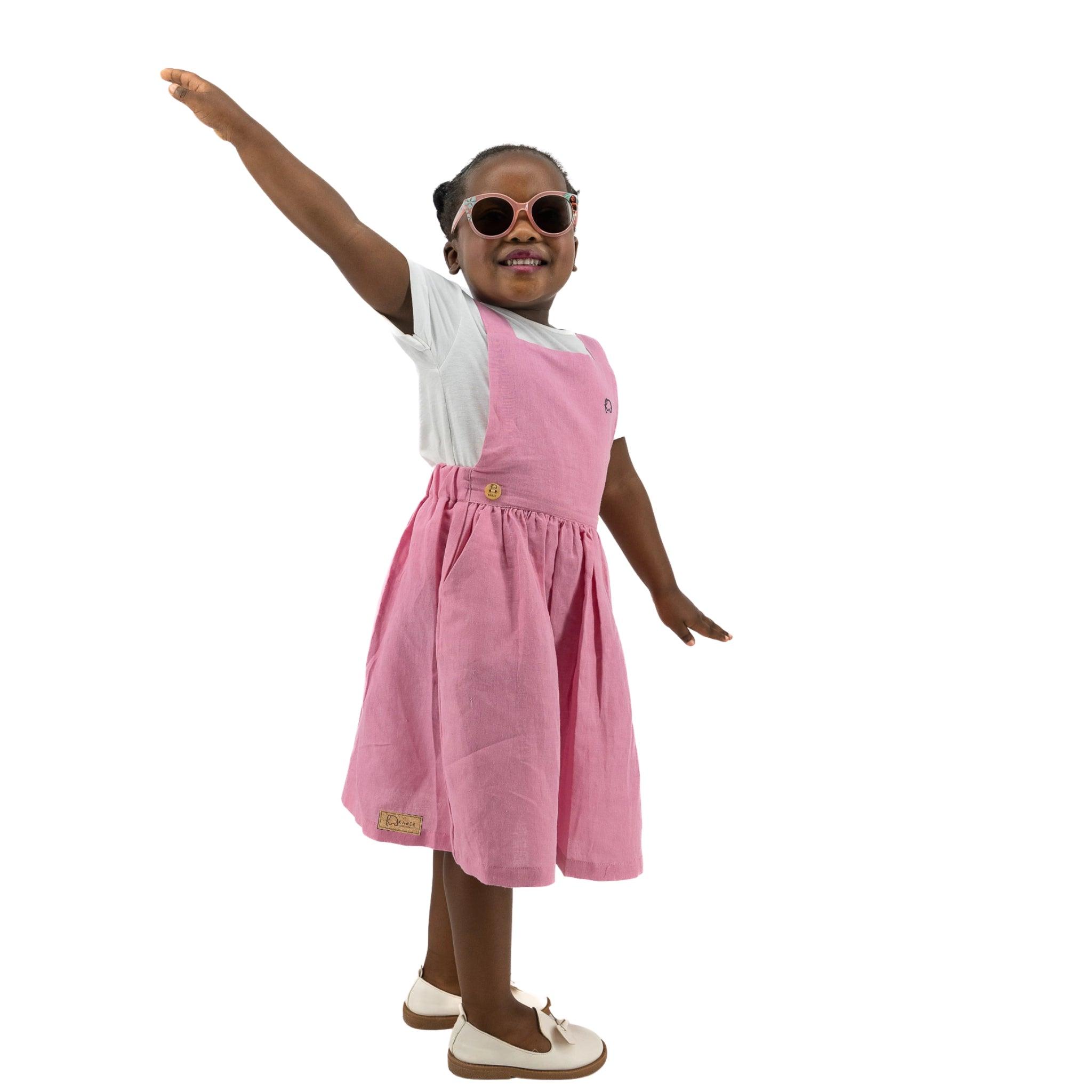 Young girl in a Karee Cashmere Rose Linen Pinafore, holding out her arm, posing playfully against a white background.