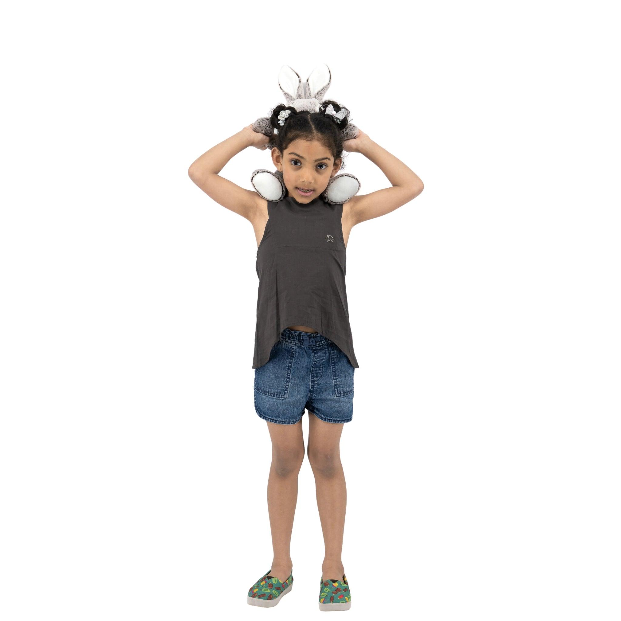A young girl wearing a Plum Kitten Cotton Bib Neck Top for Kids by Karee, denim shorts, and colorful shoes stands holding butterfly wings on her head, looking at the camera with a playful expression.