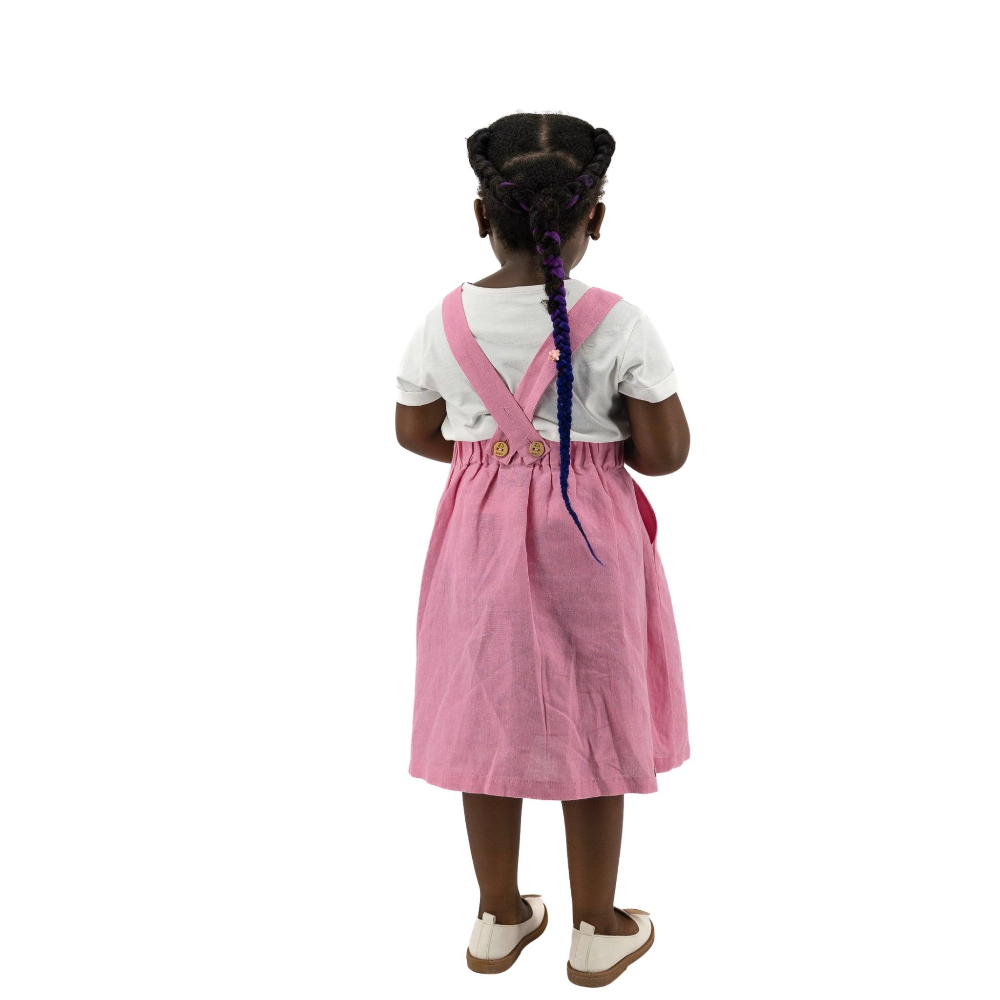 Young girl standing with her back to the camera, wearing a Karee Cashmere Rose Linen Pinafore with a white blouse, and white shoes. Her hair is styled in braids.