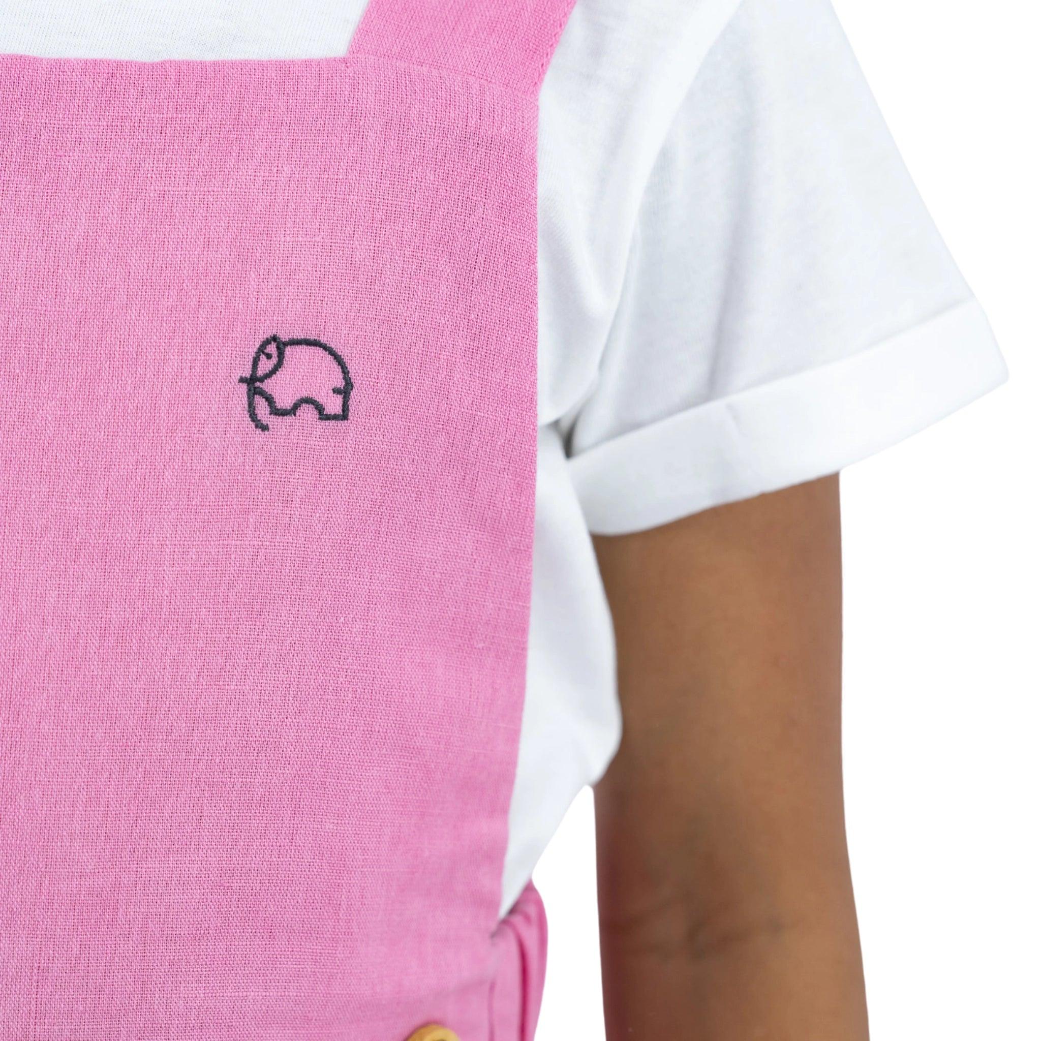 Close-up of a person wearing a Karee Cashmere Rose Linen Pinafore with a small white elephant logo over a white shirt.