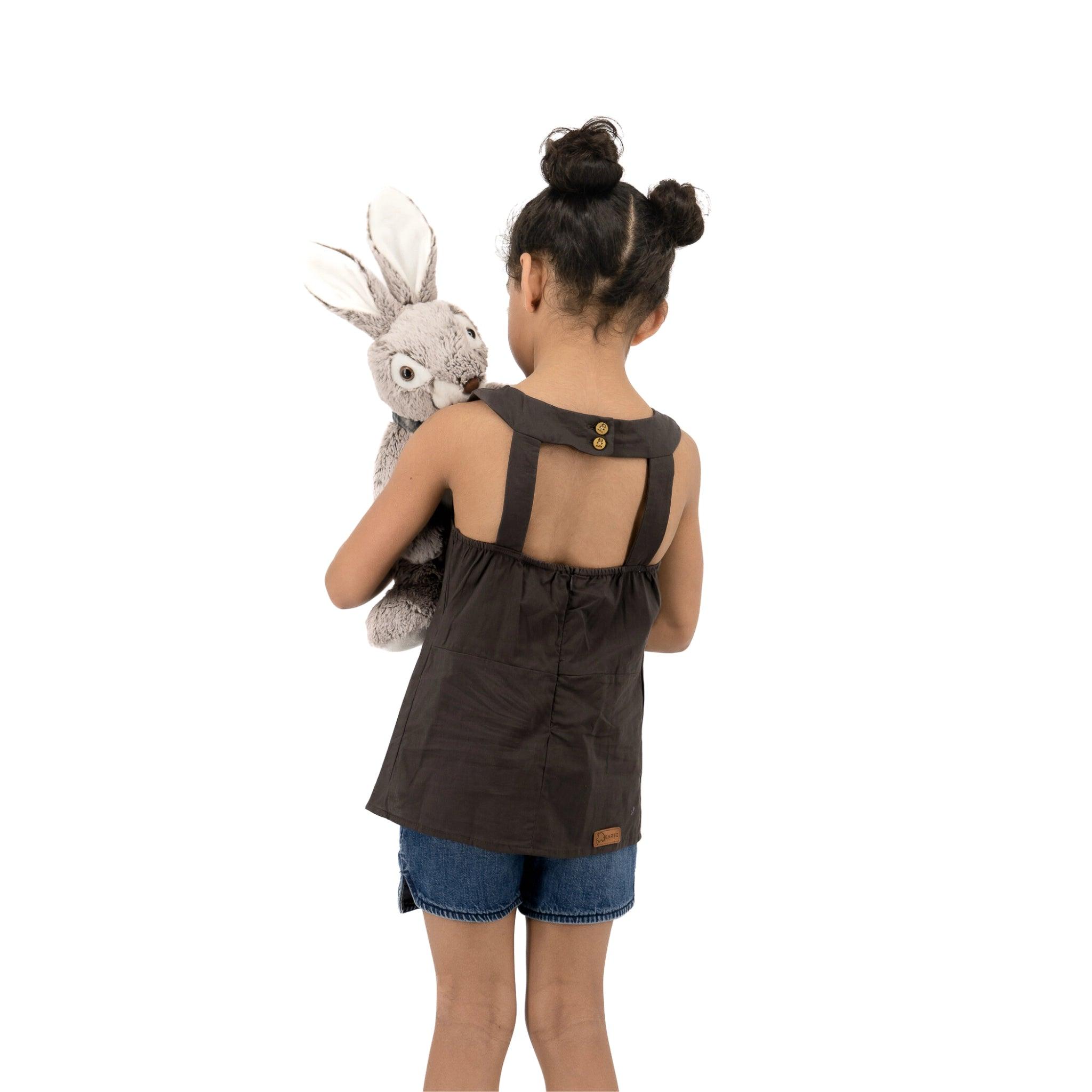 Young girl with her back to the camera, holding a stuffed rabbit, dressed in denim shorts and a Plum Kitten Cotton Bib Neck Top for Kids by Karee with hair styled in double buns.