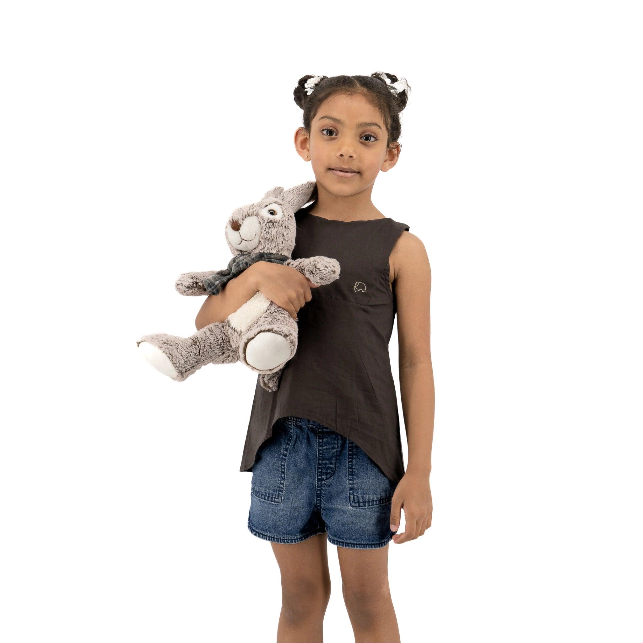 A young girl with pigtails holding a stuffed animal, wearing a Plum Kitten Cotton Bib Neck Top for Kids by Karee and denim shorts, standing against a white background.