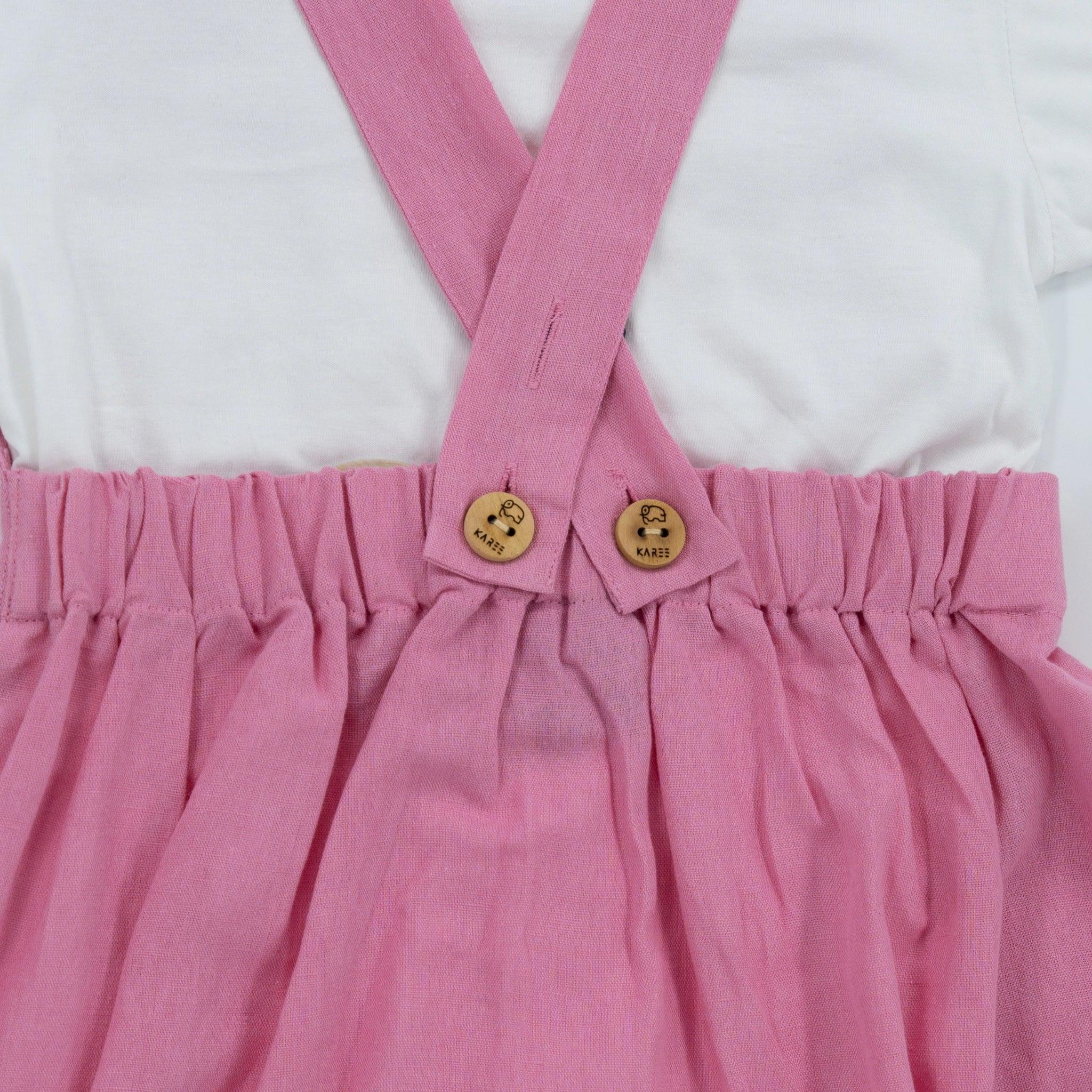 Close-up of a Karee Cashmere Rose Linen Pinafore with ruffled waist and wooden buttons attached to white suspenders.