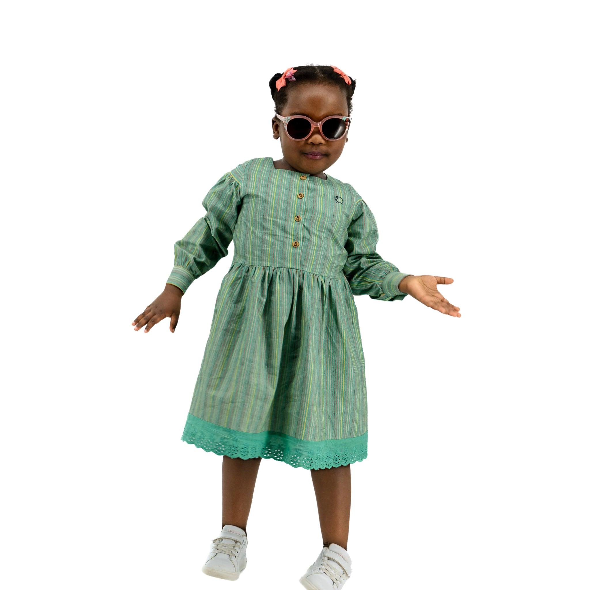 A young girl in a Karee green striped long puff sleeve cotton dress and white sneakers, wearing sunglasses and hair clips, stands smiling against a white background.