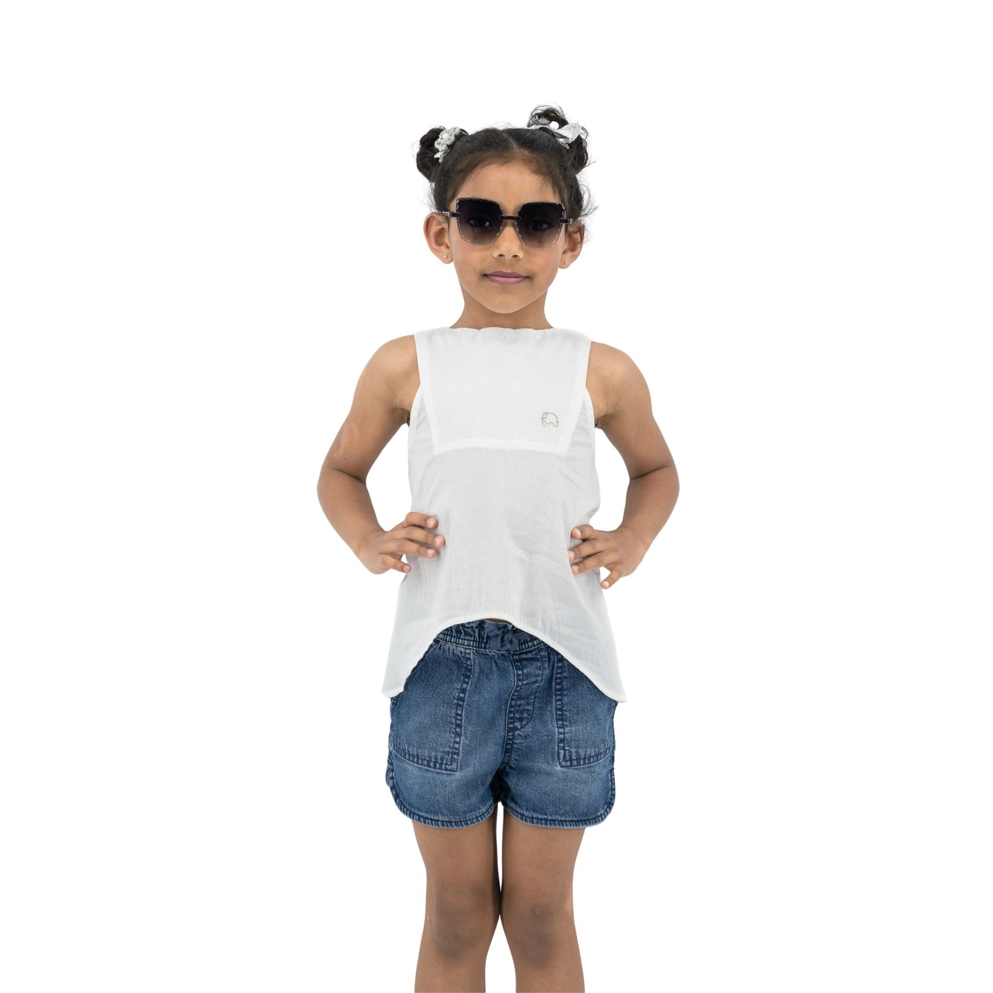 Young girl standing with hands on hips, wearing sunglasses, a Karee Brilliant White Cotton Bib Neck Top for kids and denim shorts.