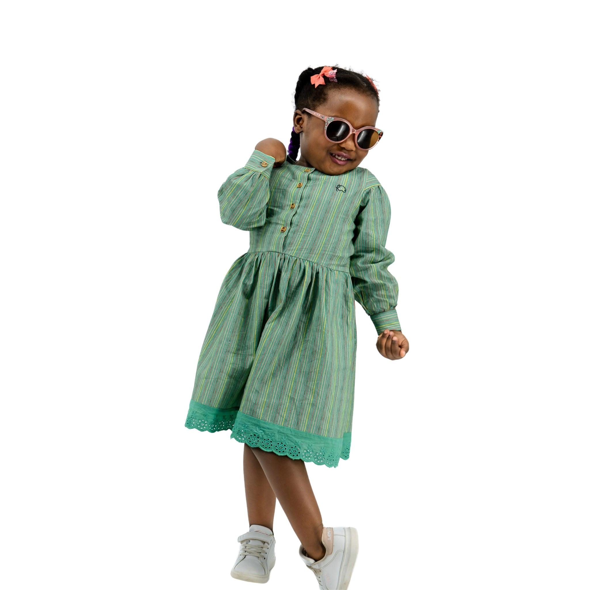 A young girl in a Karee Green Striped Long Puff Sleeve Cotton Dress and white sneakers posing playfully with sunglasses and a red hair clip, isolated on a white background.