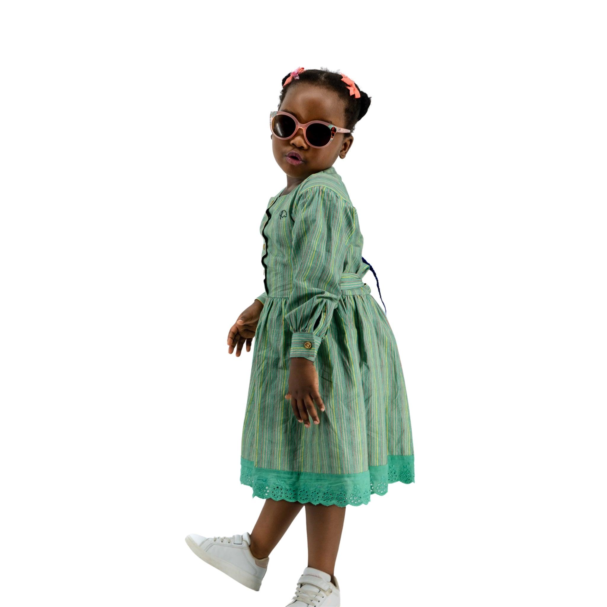 A young girl in a Karee Green Striped Long Puff Sleeve Cotton Dress and white sneakers, wearing sunglasses and hair clips, standing confidently against a white background.