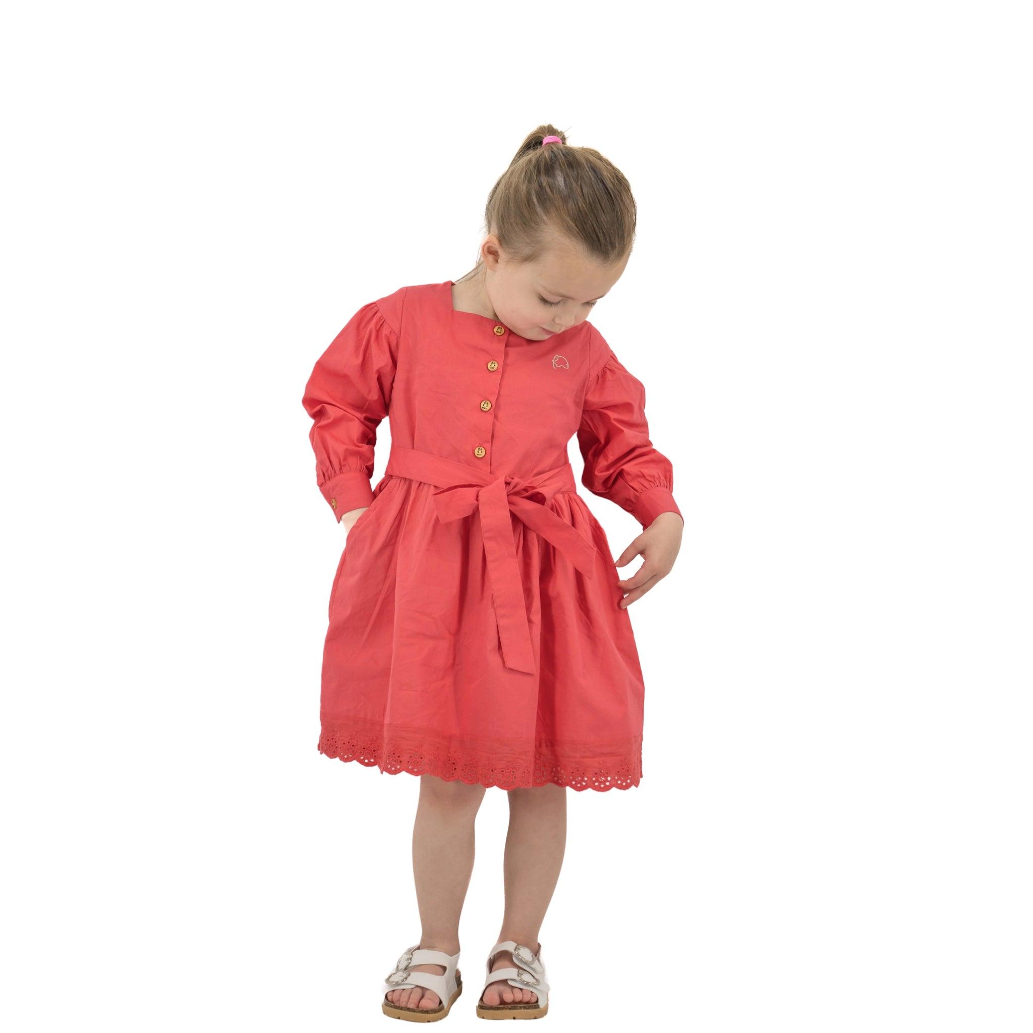 A young girl in a Karee Red Long Puff Sleeve Cotton Dress looking down at her dress, standing against a white background.