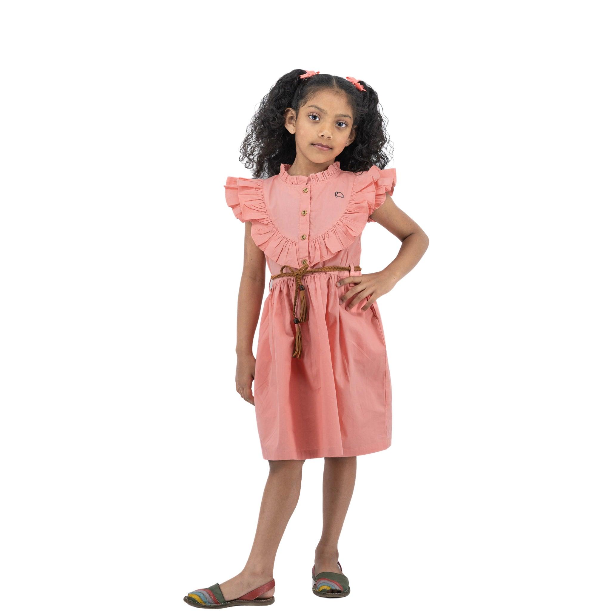 Young girl with curly hair, wearing a Karee peach and cream butterfly sleeve cotton dress and multicolored sandals, standing and looking thoughtfully at the camera on a white background.