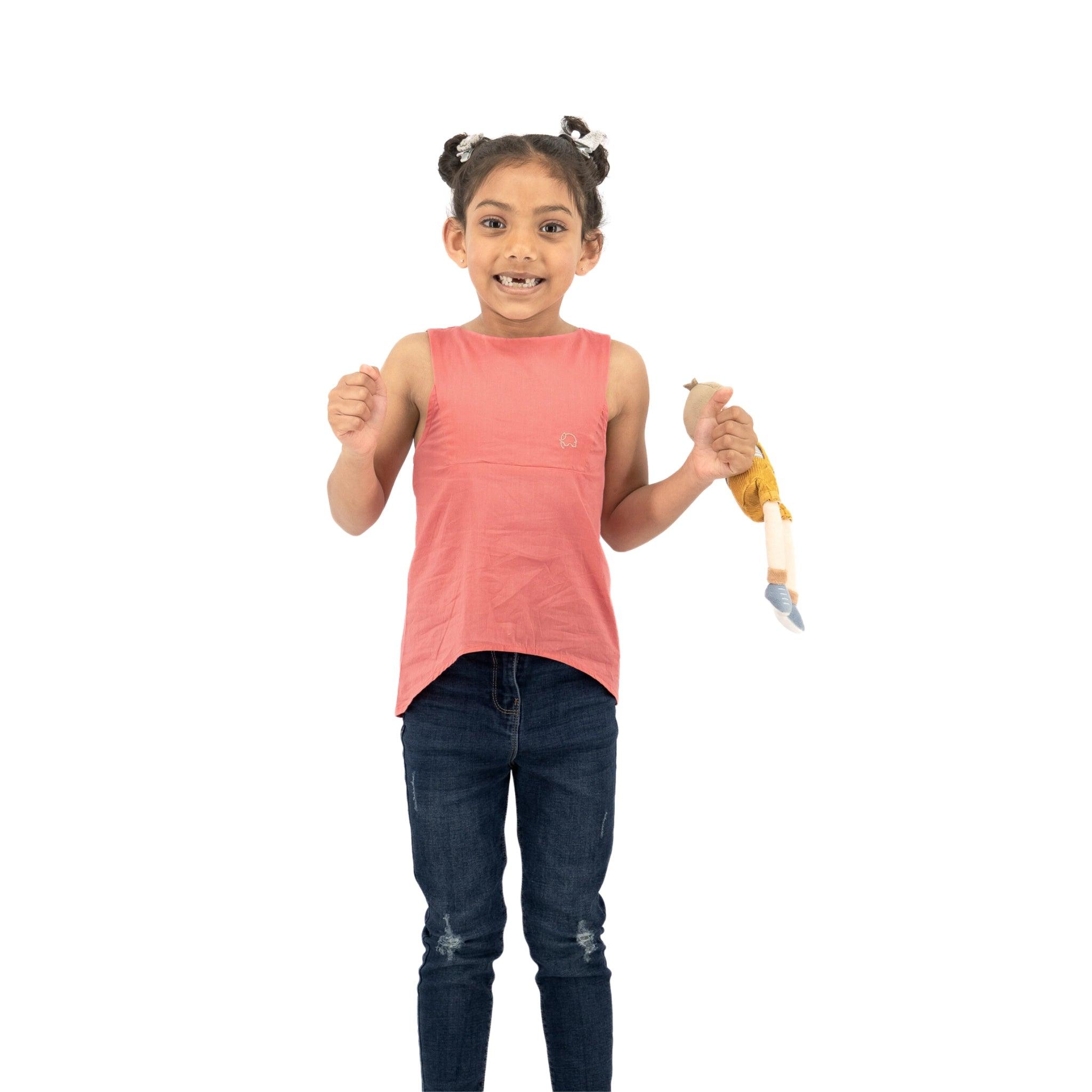 A young girl with a playful expression, wearing a Karee Tea Rose Cotton Bib Neck Top for kids, holds a banana with a toy parachute against a white background.