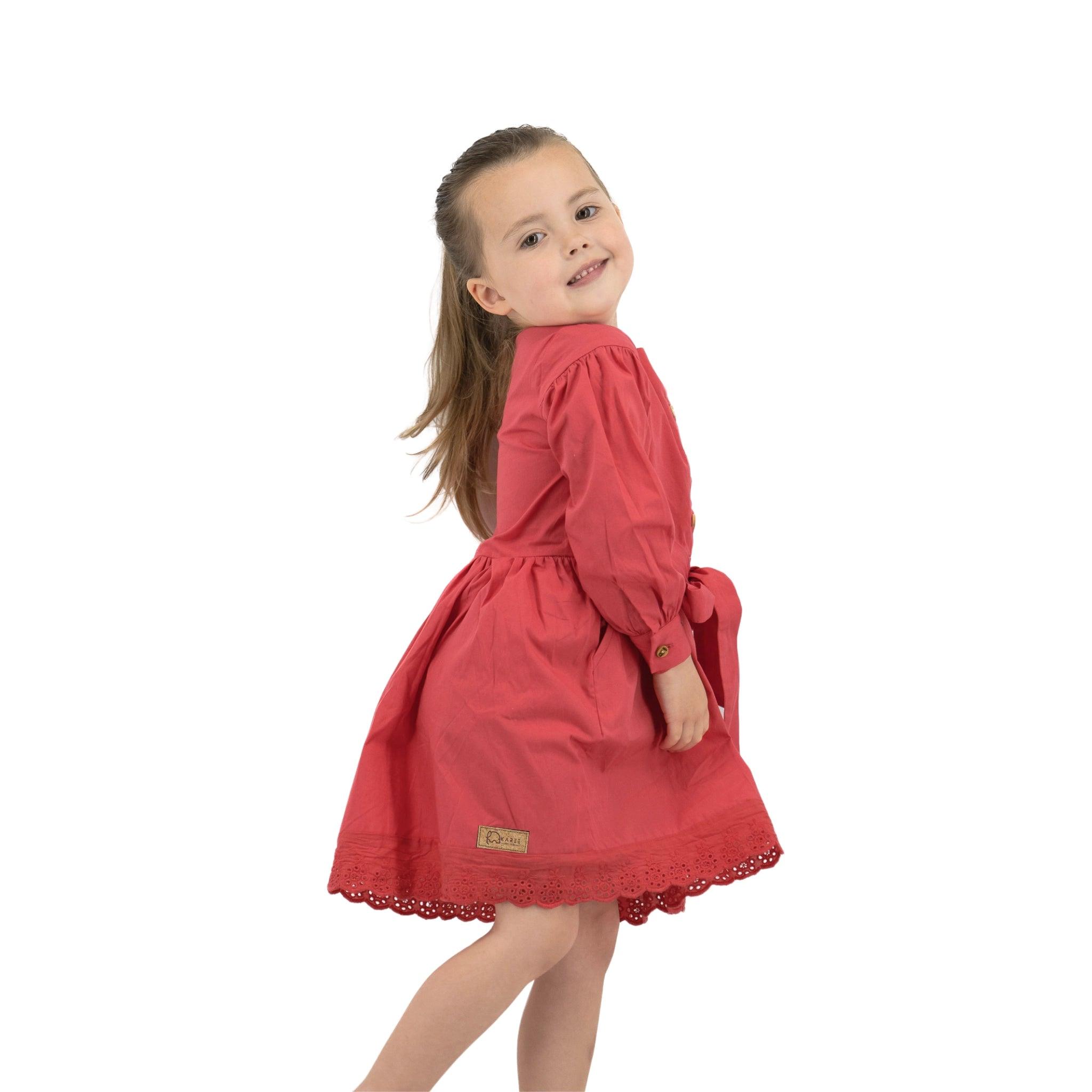 Young girl in a Karee red long puff sleeve cotton dress playfully twirling, looking over her shoulder with a smile, isolated on a white background.