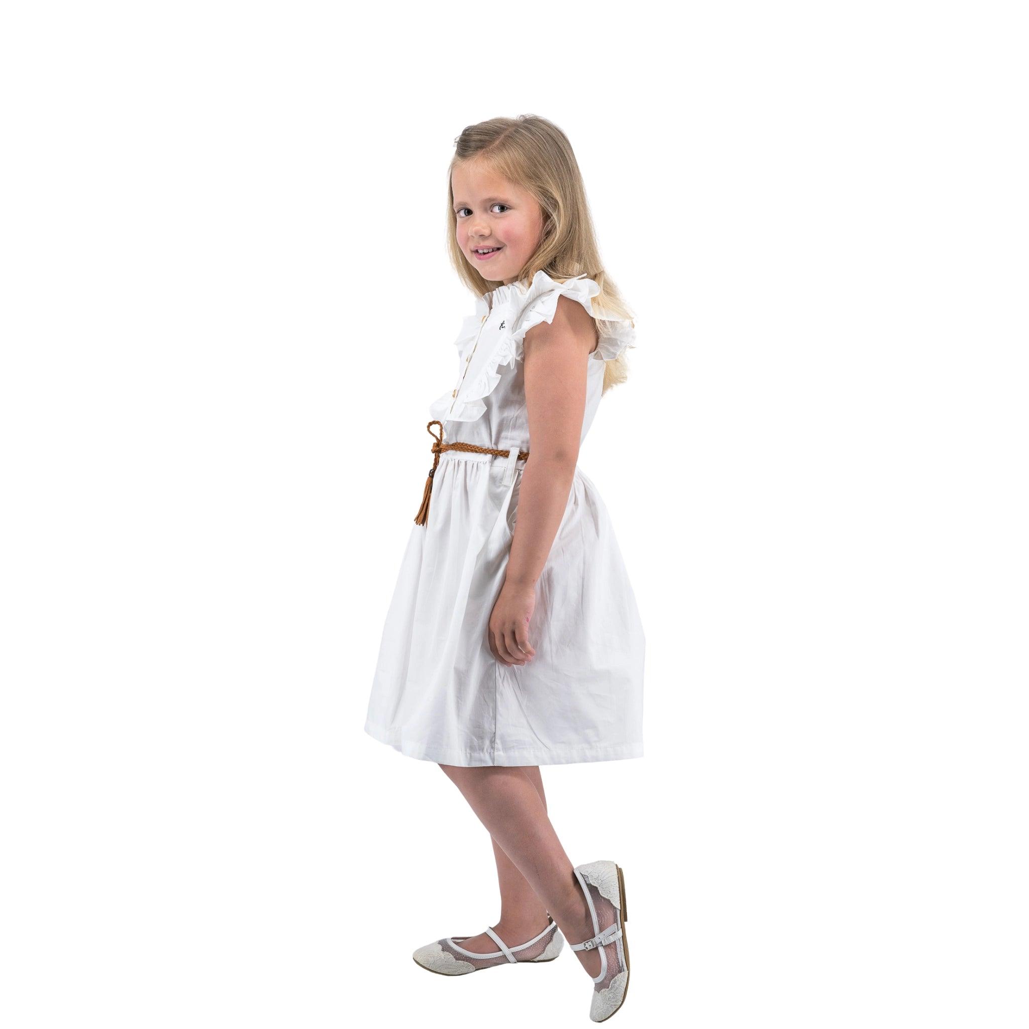 Young girl in a Karee Butterfly Sleeve Cotton Dress in White and silver shoes, smiling over her shoulder against a white background.
