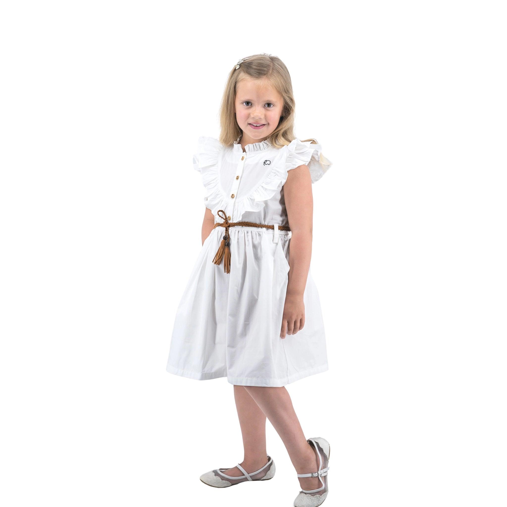 A young girl in a Karee Butterfly Sleeve Cotton Dress in White with a brown belt and silver shoes, standing and smiling against a white background.