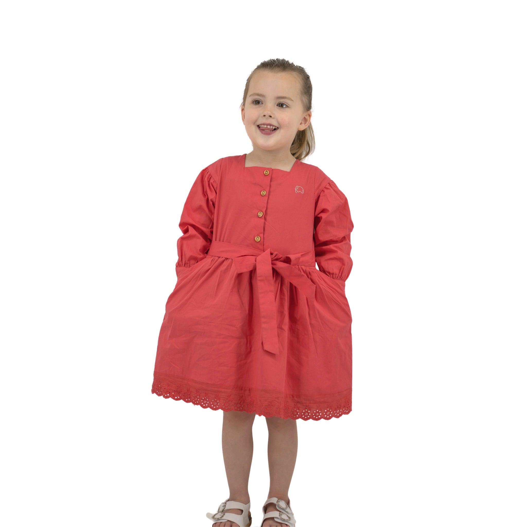 Young girl smiling in a Karee red long puff sleeve cotton dress with buttons and a bow, standing against a white background.