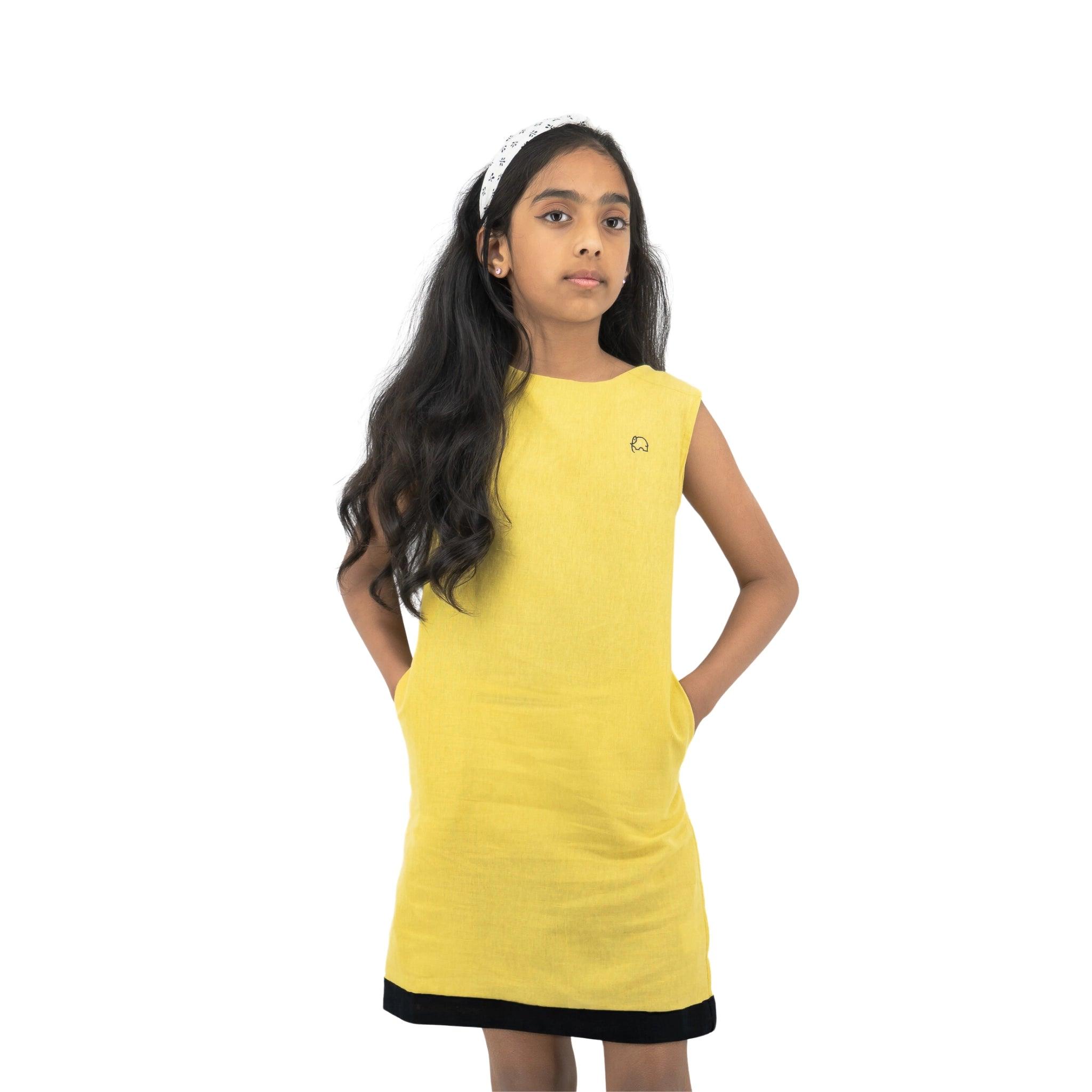 A young girl wearing a Karee cream gold Linen Cotton Round Neck Frock for Kids and a white headband stands with her hands on her hips, looking to the side.