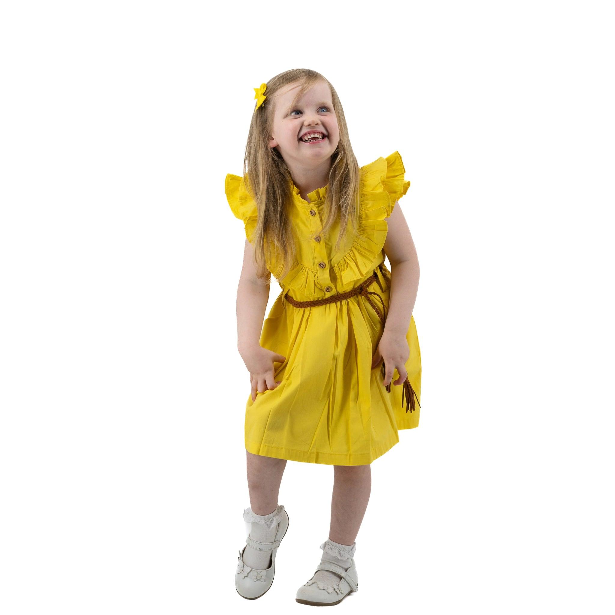 A young girl in a Karee yellow butterfly sleeve cotton dress and white shoes smiles joyfully, looking upwards, isolated on a white background.