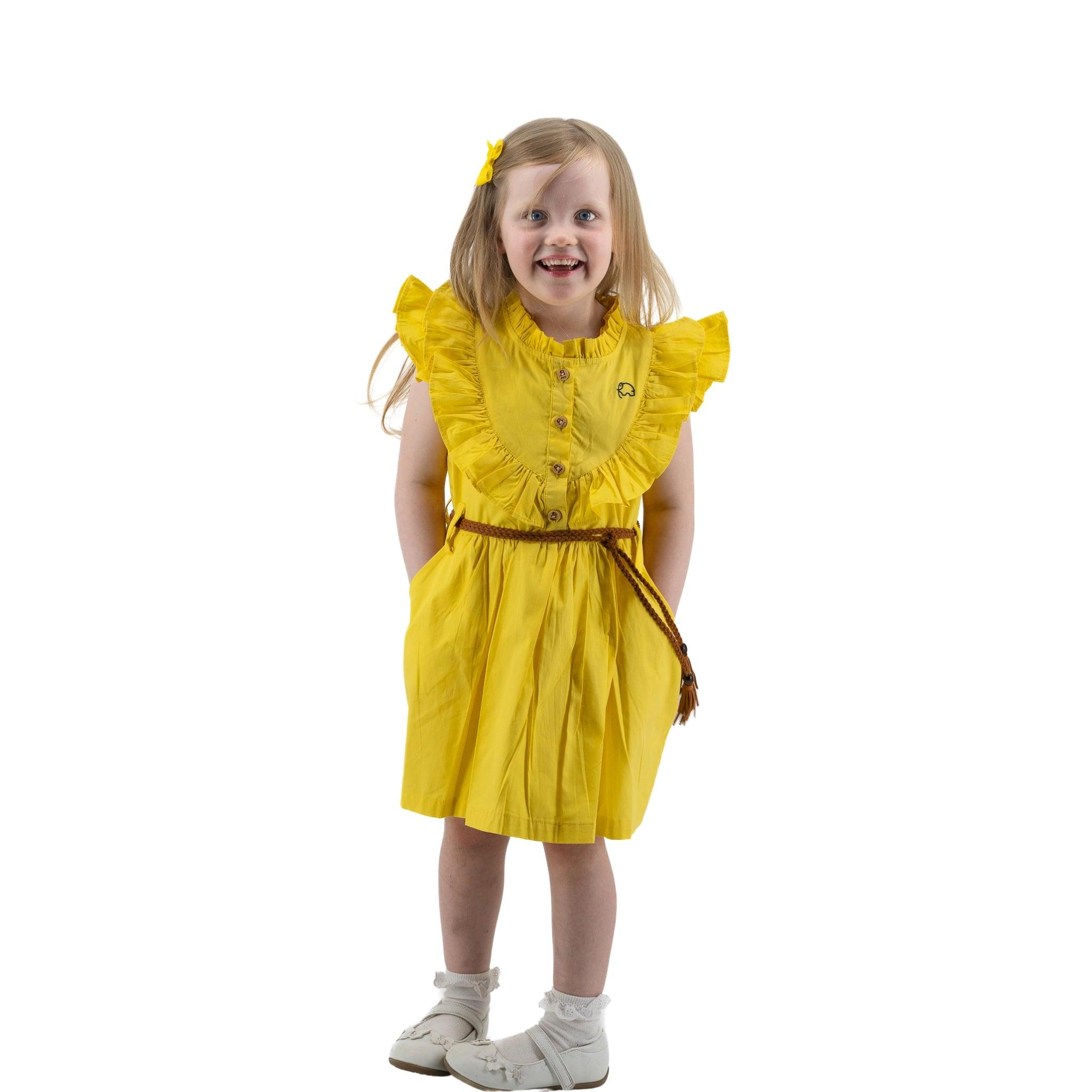 A young girl wearing a Karee yellow butterfly sleeve cotton dress for girls and white shoes smiles while posing with her hands on her hips, isolated on a white background.