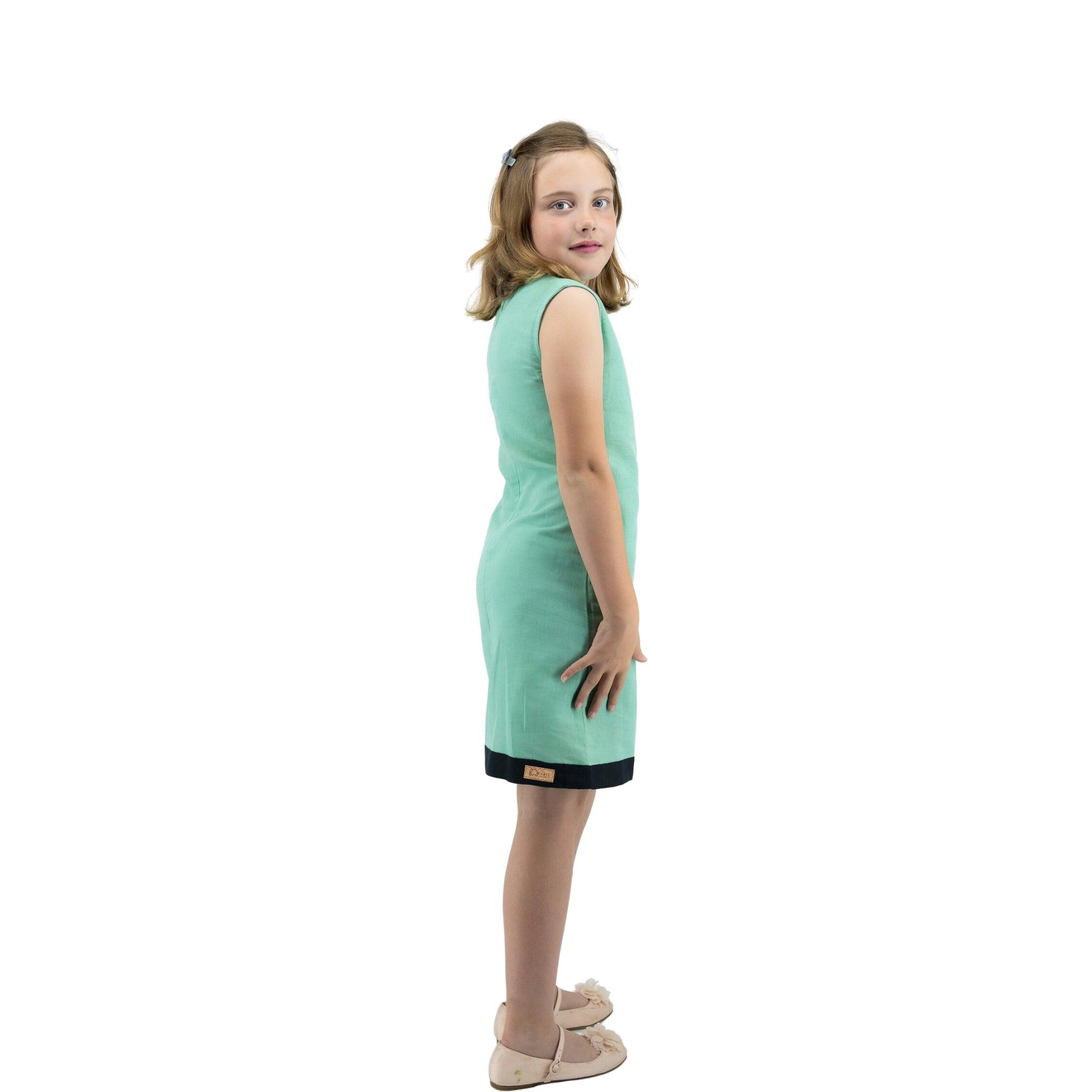 Young girl in a Karee Linen Cotton Round Neck Frock for Kids in Neptune Green and beige shoes, looking over her shoulder with a slight smile, standing against a white background.
