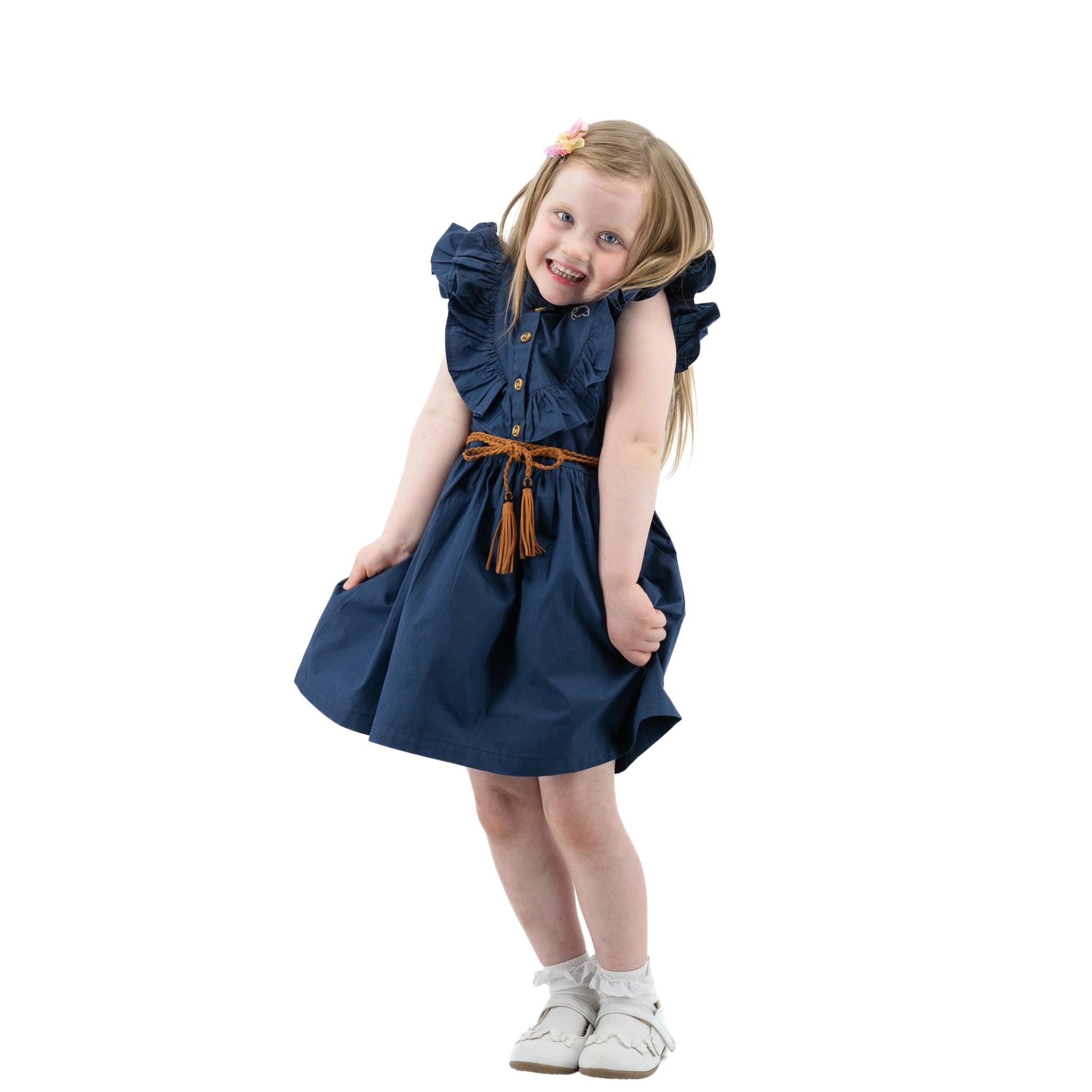 A young girl in a Karee navy blue butterfly sleeve cotton dress playfully tilting her head, holding her dress with one hand and standing isolated on a white background.