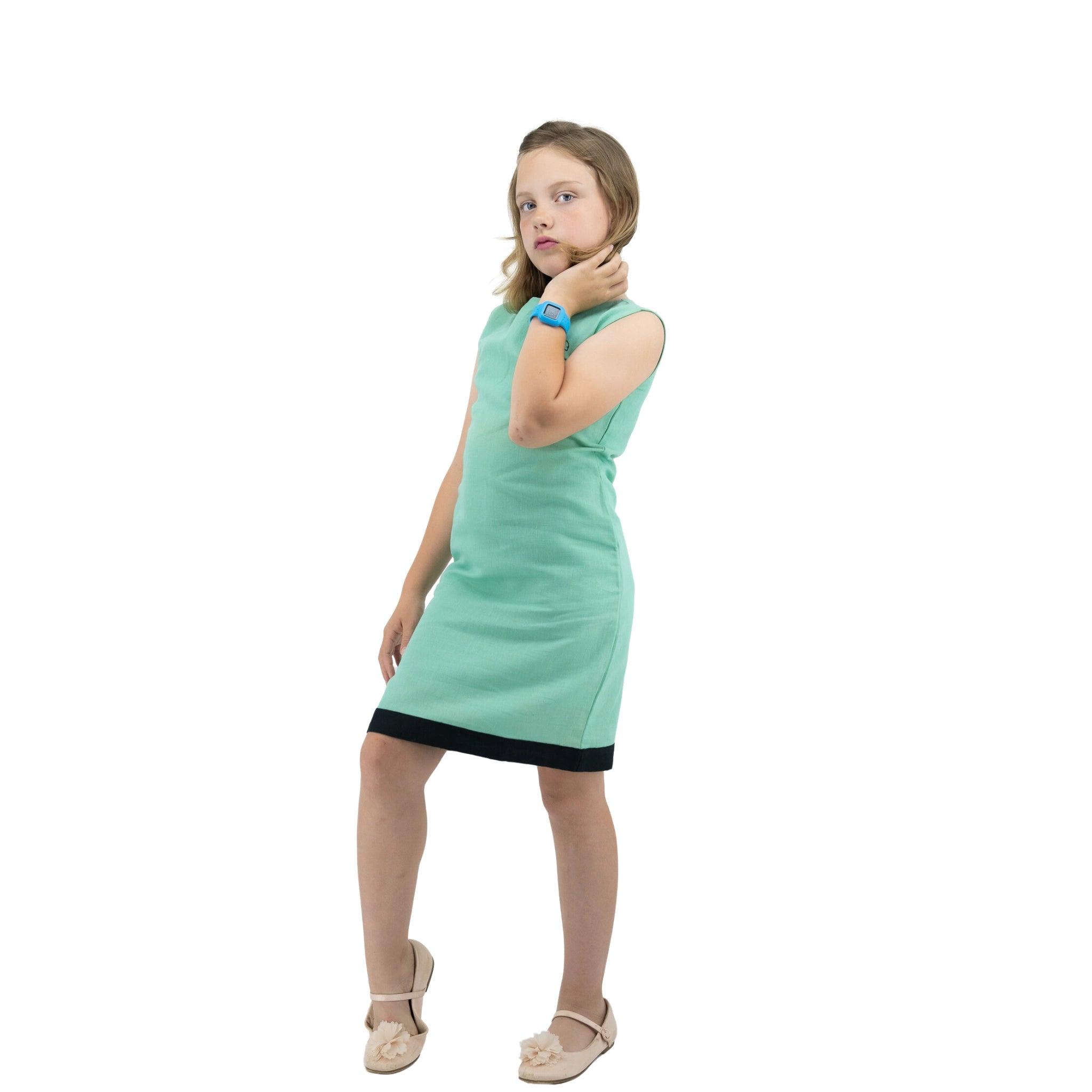 A young girl standing in a thoughtful pose, wearing a Karee Linen Cotton Round Neck Frock for Kids in Neptune Green with a black hemline and light-colored flats, isolated on a white background.