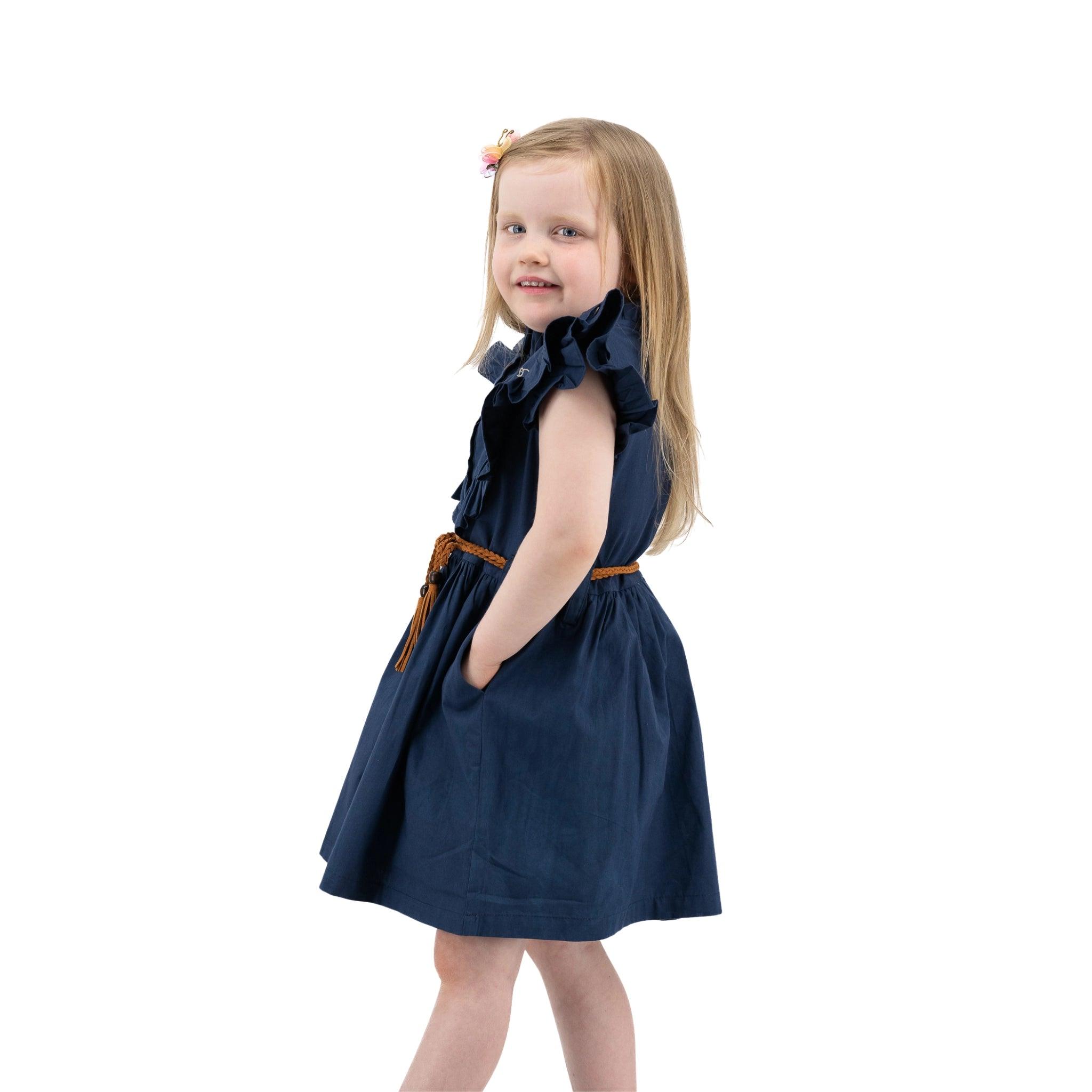 A young girl in a Karee Navy Blue Butterfly Sleeve Cotton Dress smiling and looking over her shoulder, standing against a white background.