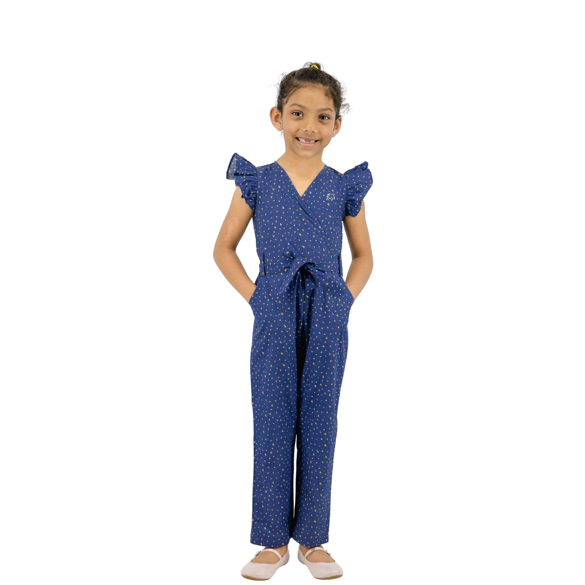 Young girl in a Karee Estate Blue Cotton V-Neck jumpsuit standing with hands behind back, smiling at the camera on a white background.