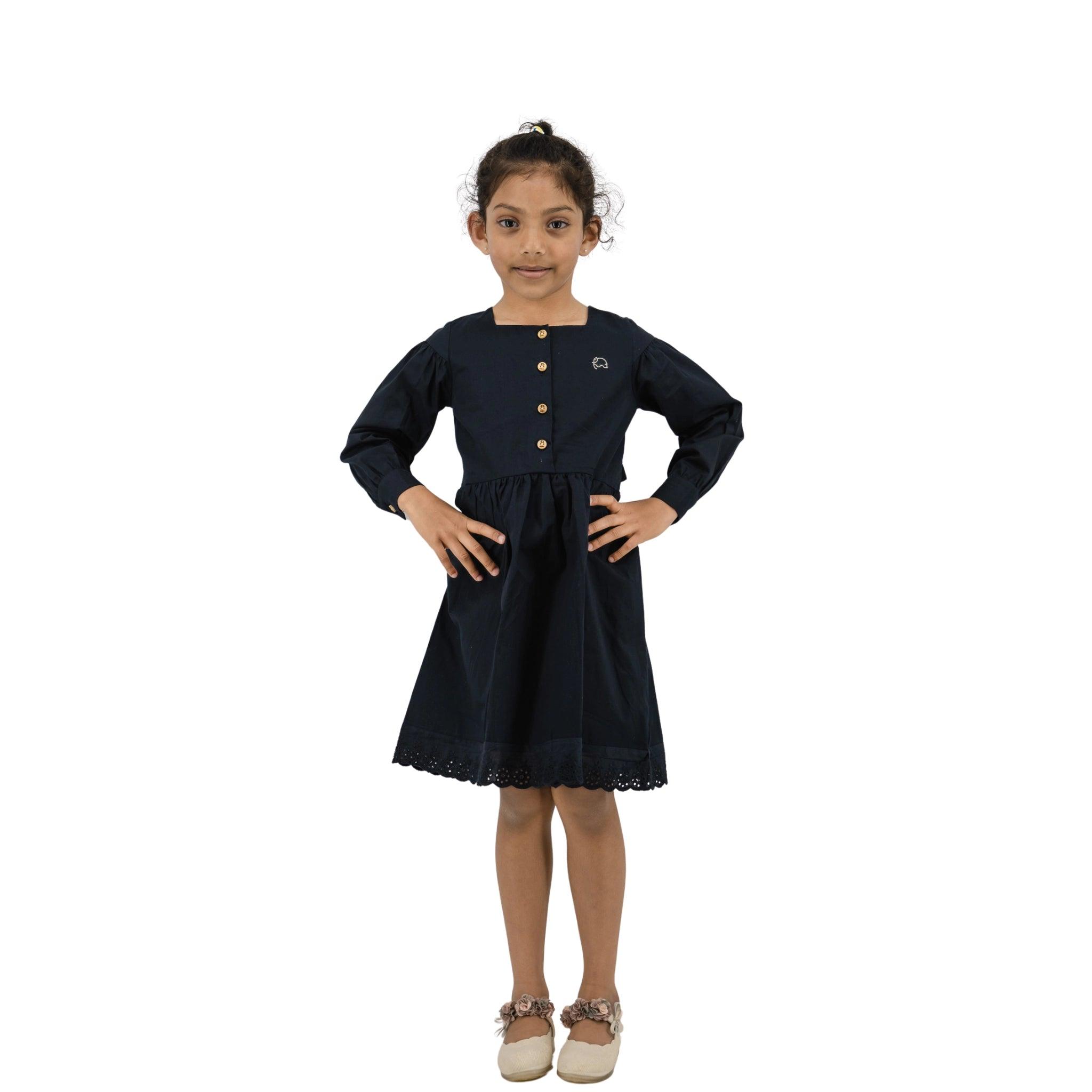A girl in a Karee black long puff sleeve cotton dress with white shoes, standing with hands on her hips, smiling, against a white background.