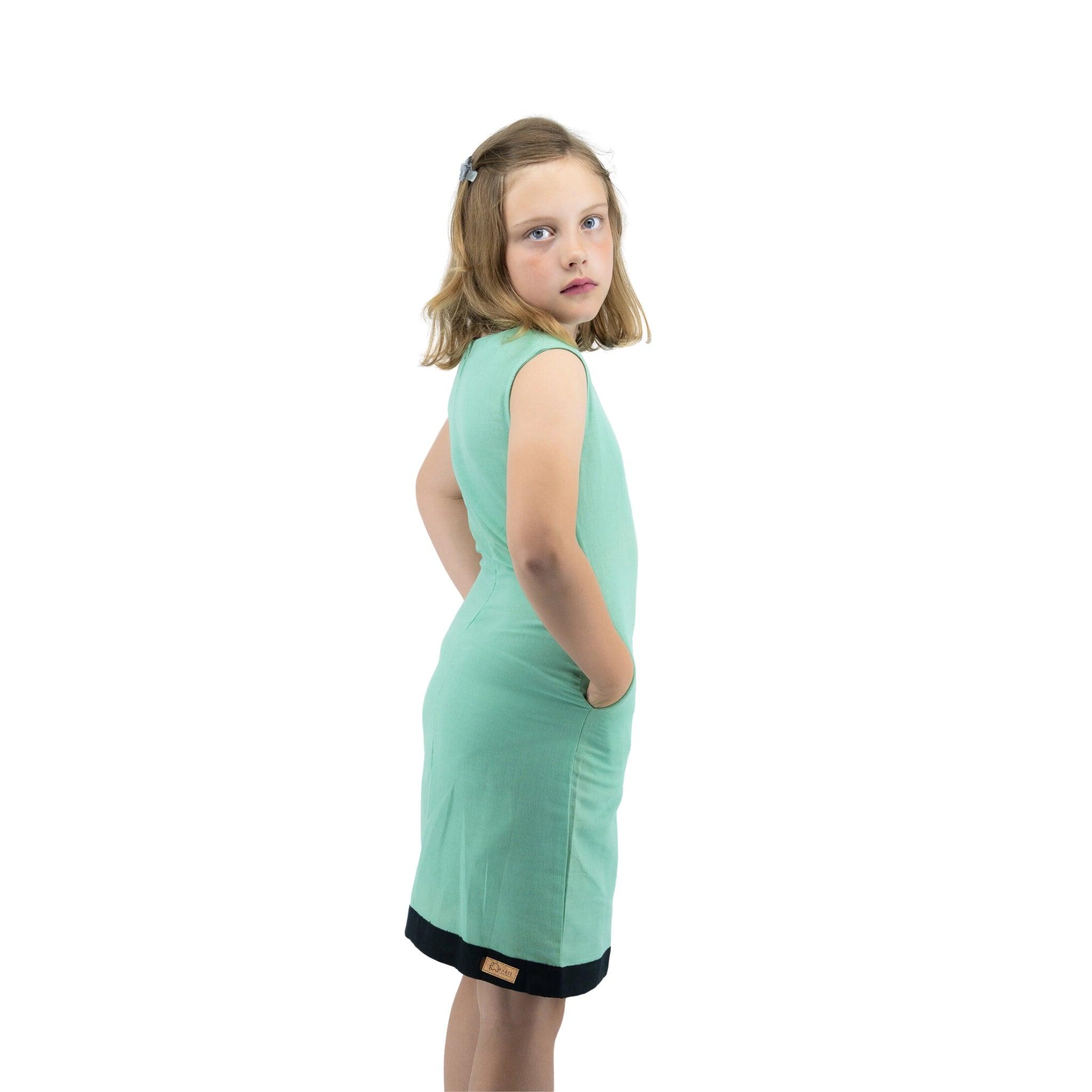 Young girl in a Karee Linen Cotton Round Neck Frock for Kids in Neptune Green looking over her shoulder, standing against a white background.