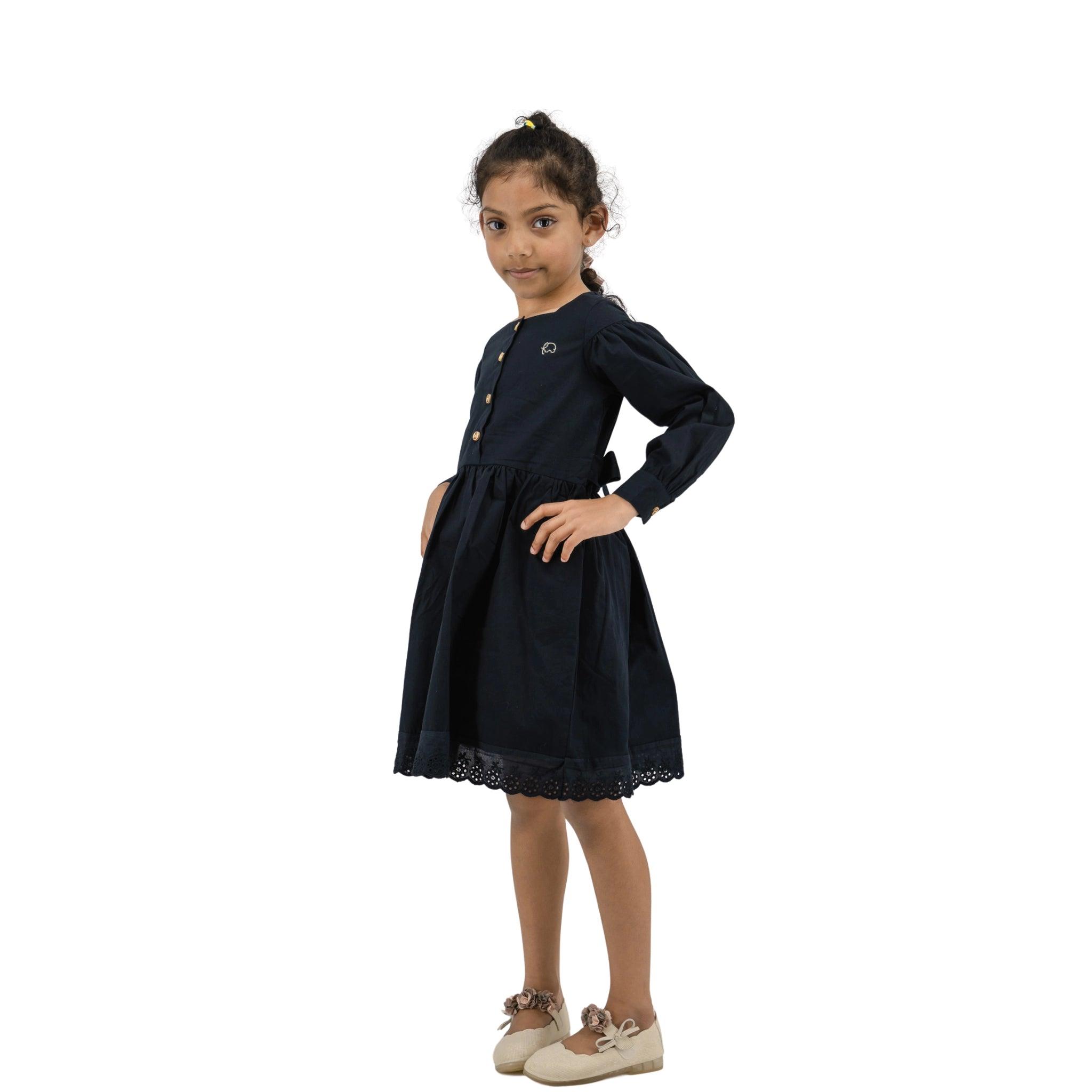 Young girl posing in a navy blue Karee dress with long puff sleeves and lace detailing, hands on hips, standing against a white background.
