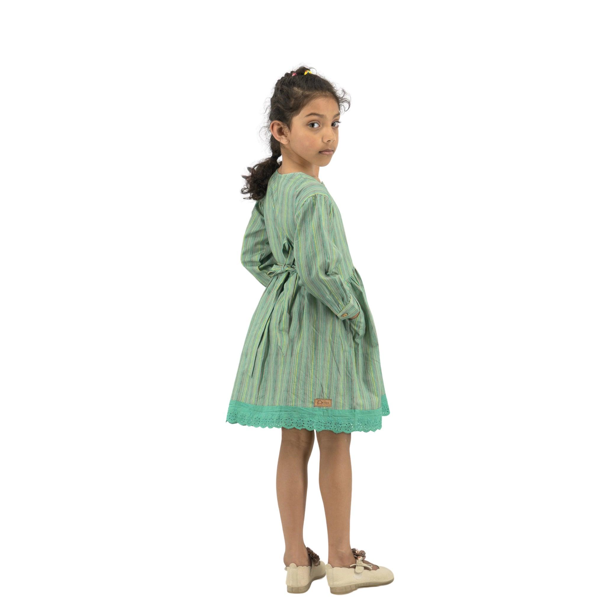 Young girl in a Karee Green Striped Long Puff Sleeve Cotton Dress and white shoes, standing sideways, looking over her shoulder with a curious expression.