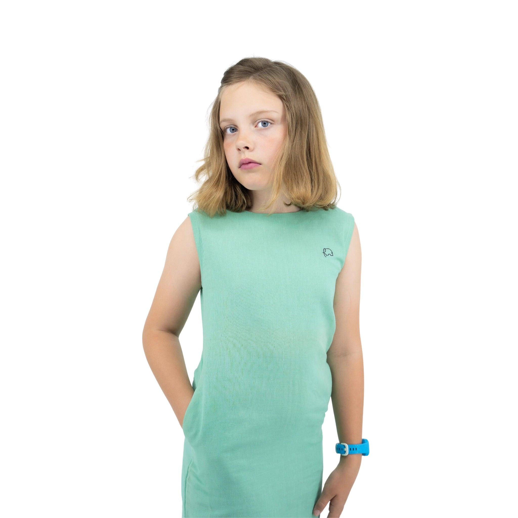 Young girl in a Karee Linen Cotton Round Neck Frock for Kids in Neptune Green standing against a white background, looking slightly to the side with a thoughtful expression.