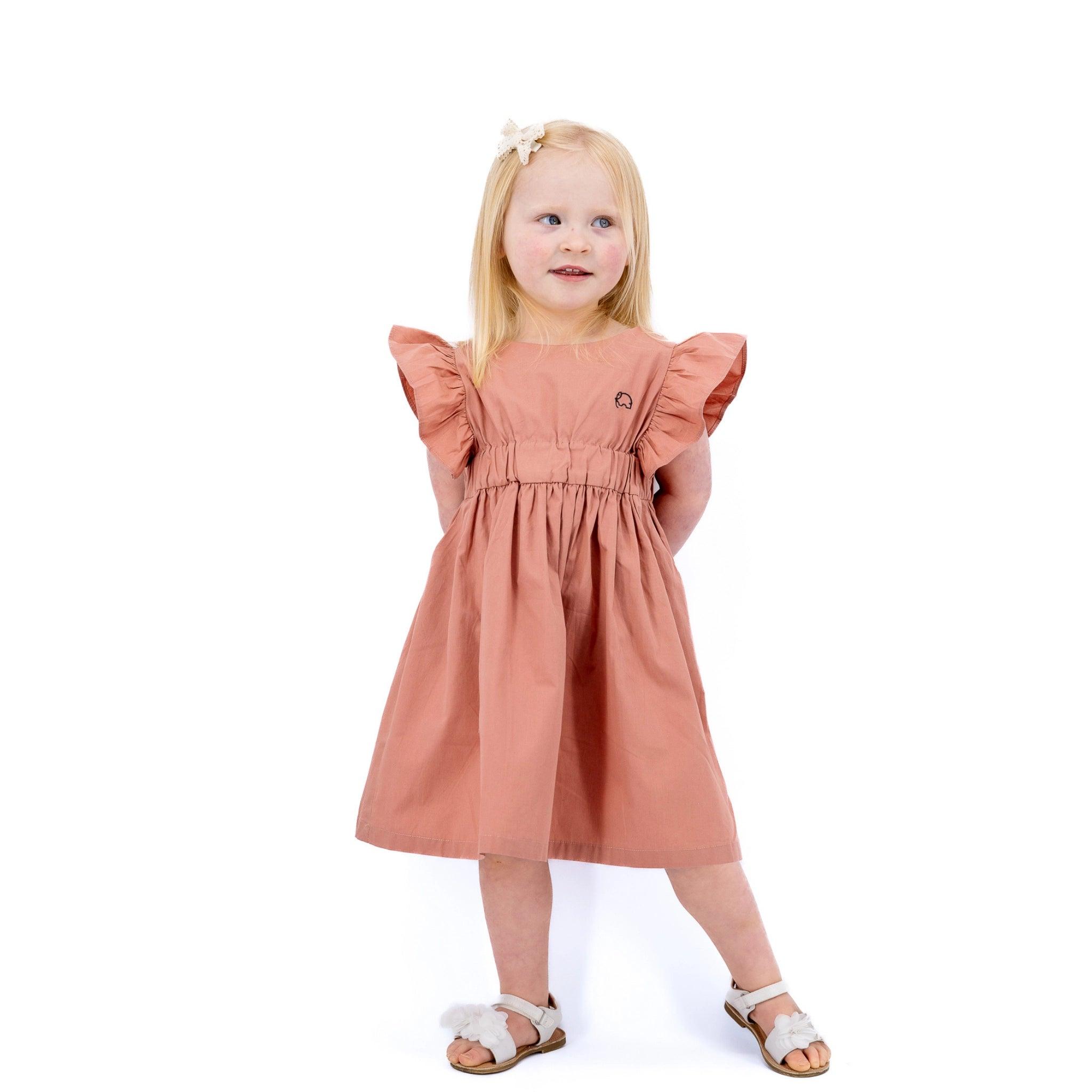 Young girl with blonde hair, wearing a Karee Brick Dust Cotton Floral Dress for Girls and white sandals, standing and smiling against a white background.